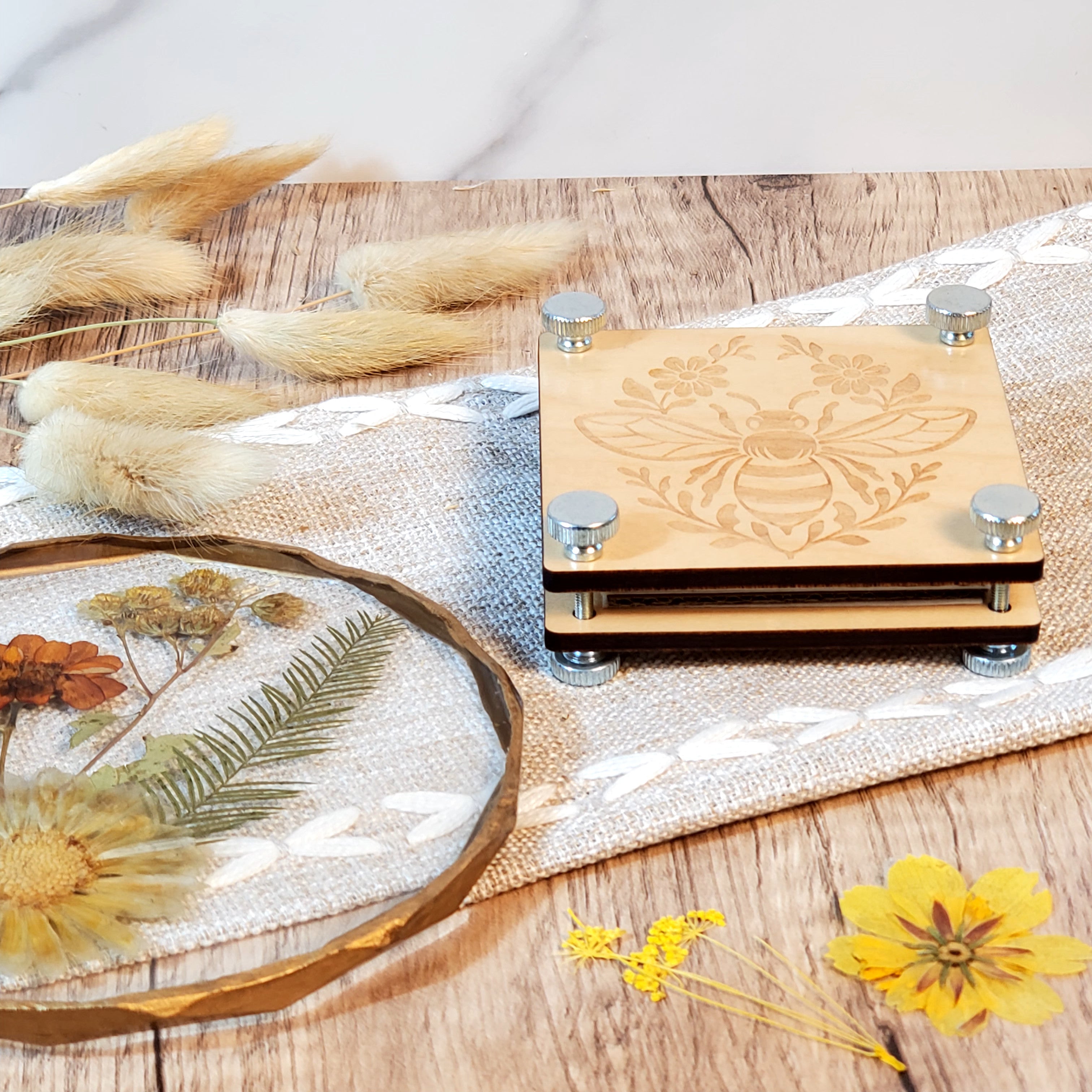 Laser engraved wooden flower press with a bee and floral design, shown with metal bolts and styled with dried flowers on a rustic wood surface.