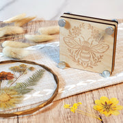 Laser engraved wooden flower press with a bee and floral design, shown with metal bolts and styled with dried flowers on a rustic wood surface.