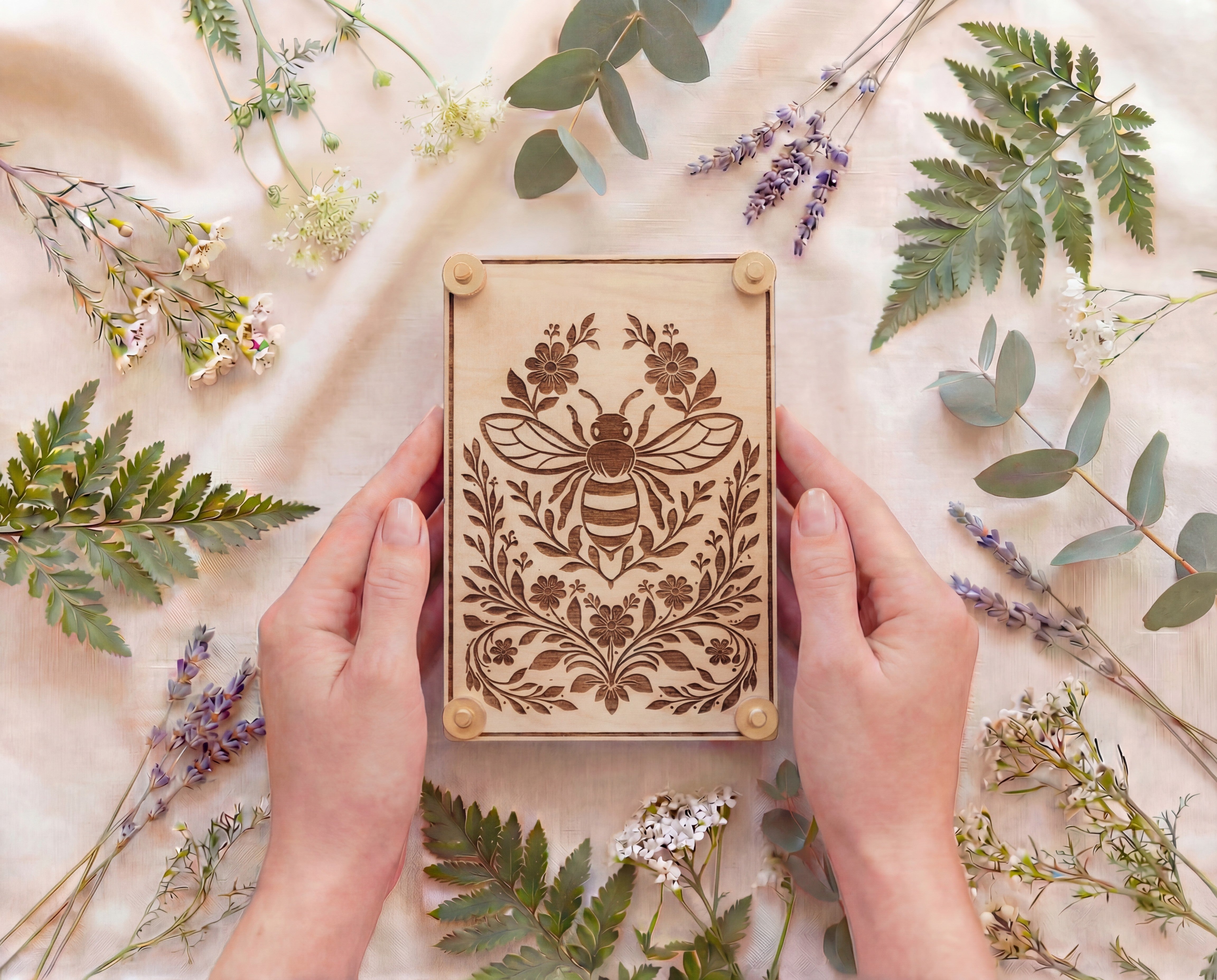 Hands holding a 4x6 engraved wooden flower press kit with a bee and botanical floral design, made from 5.2mm premium maple MDF core board with brass hardware and knurled thumb nuts, photographed overhead on a linen background surrounded by fresh greenery and flowers for pressing and preserving botanicals.