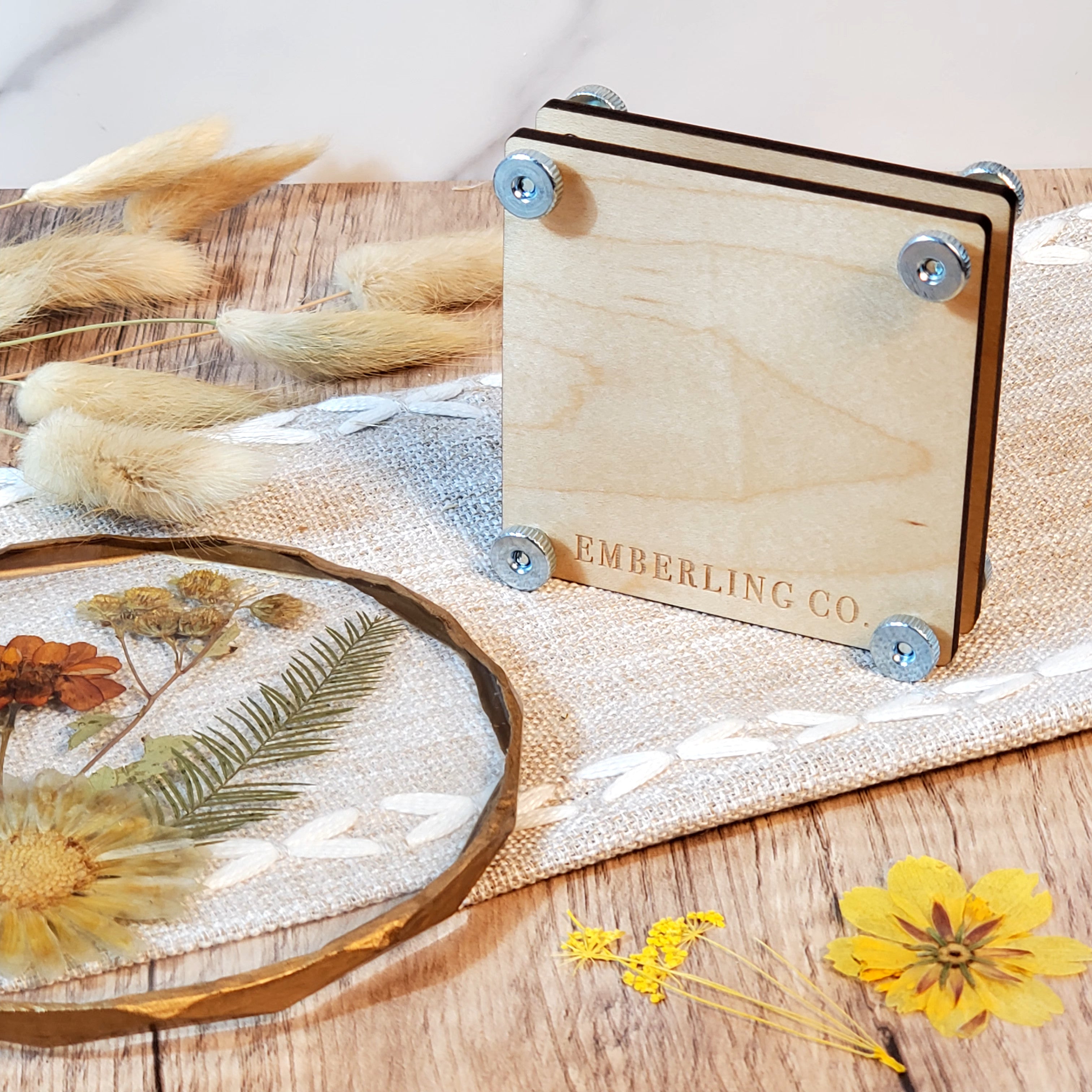 Laser engraved wooden flower press with a fern and floral design, shown with metal bolts and styled with dried flowers on a rustic wood surface.