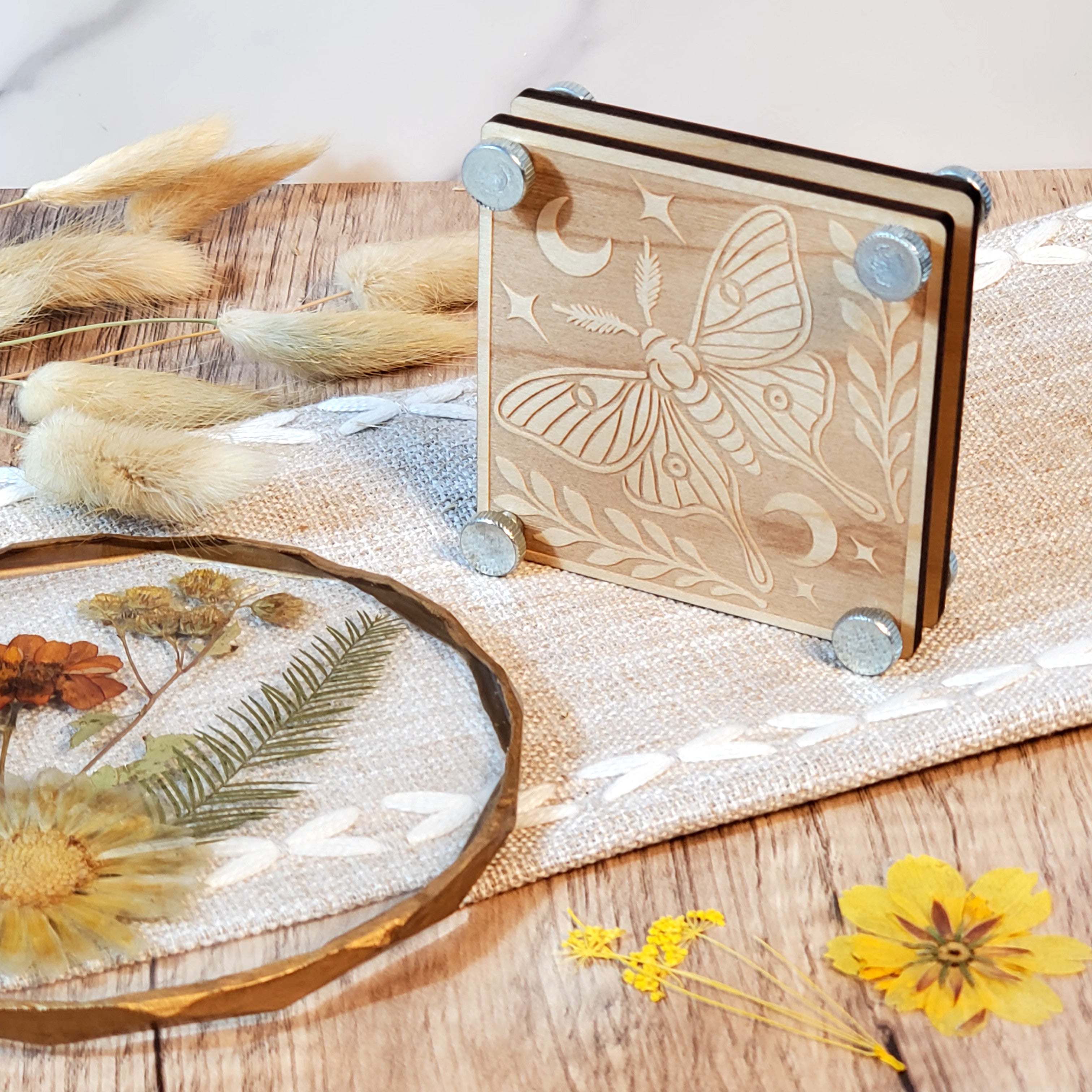 Laser engraved wooden flower press with a celestial moth design, shown with metal bolts and styled with dried flowers on a rustic wood surface.