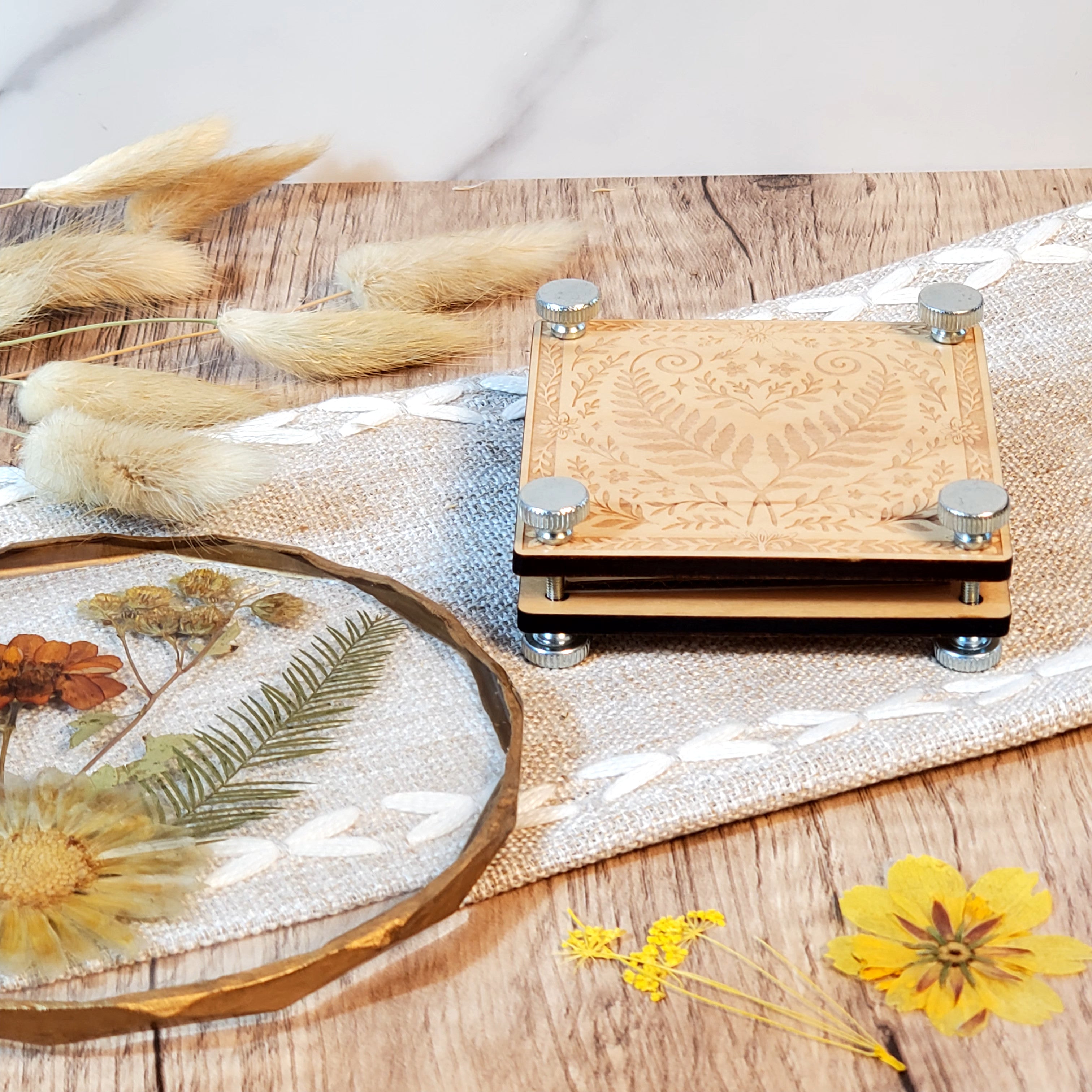 Laser engraved wooden flower press with a fern and floral design, shown with metal bolts and styled with dried flowers on a rustic wood surface.