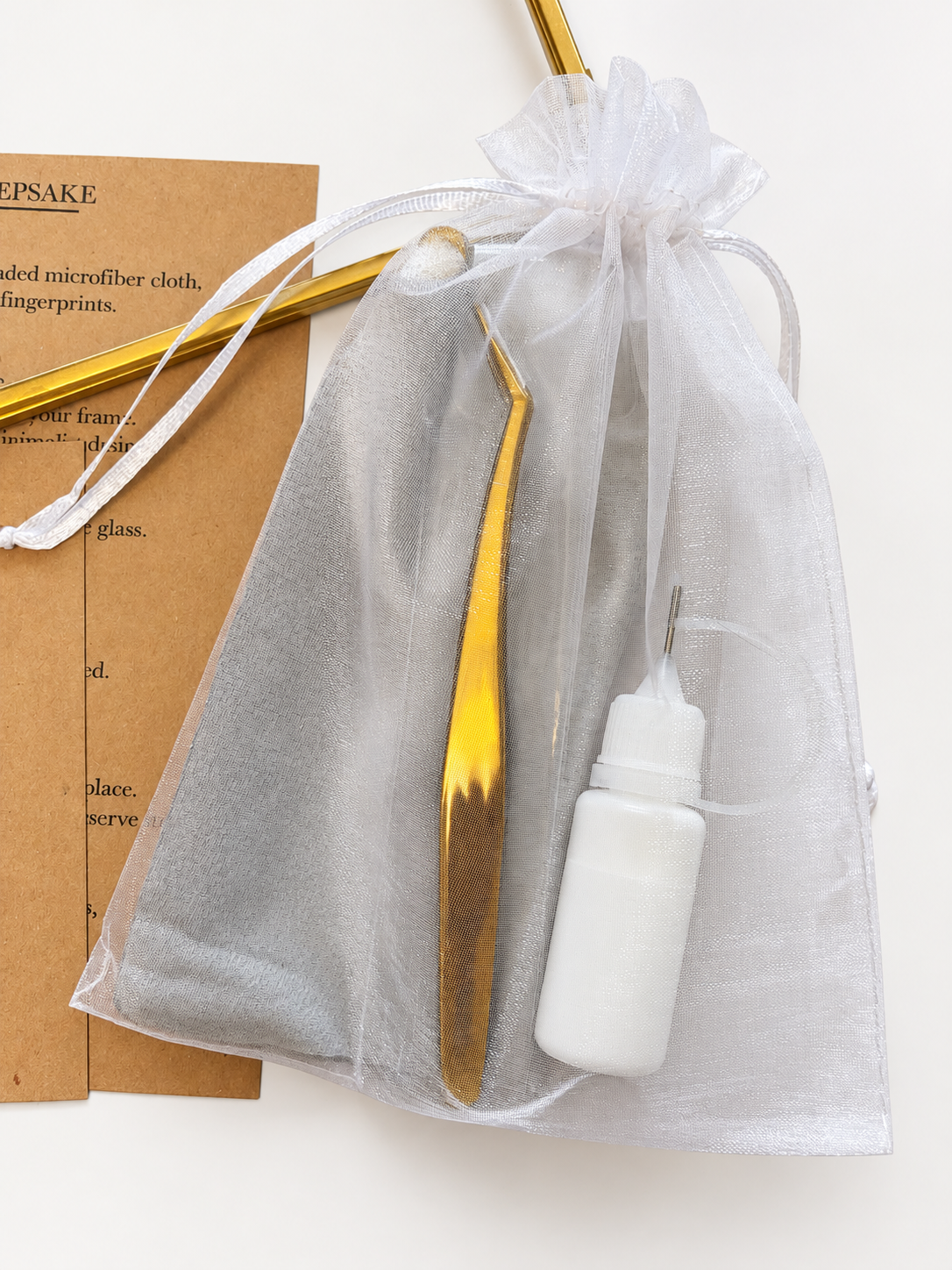 Gold precision tweezers, shown inside a sheer white organza drawstring pouch alongside a small white bottle filled with glue and a brown kraft paper instruction card.