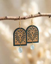 Wooden green painted arch shaped earrings with leaf patterns and light blue teardrop Czech glass bead hanging from a branch.