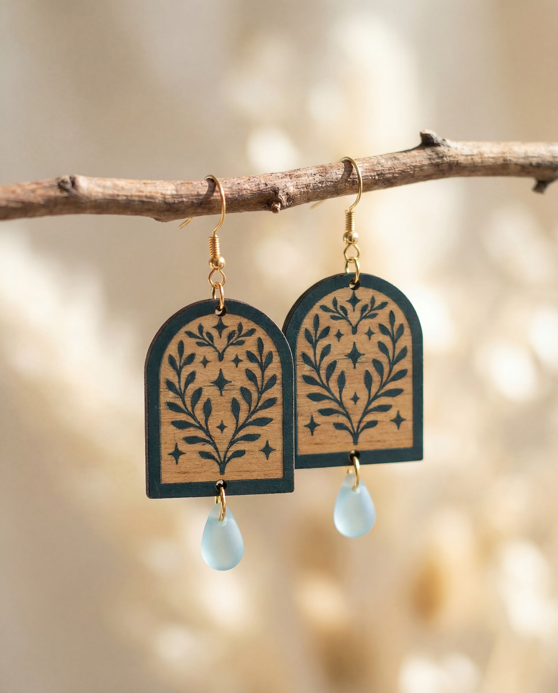 Wooden green painted arch shaped earrings with leaf patterns and light blue teardrop Czech glass bead hanging from a branch.