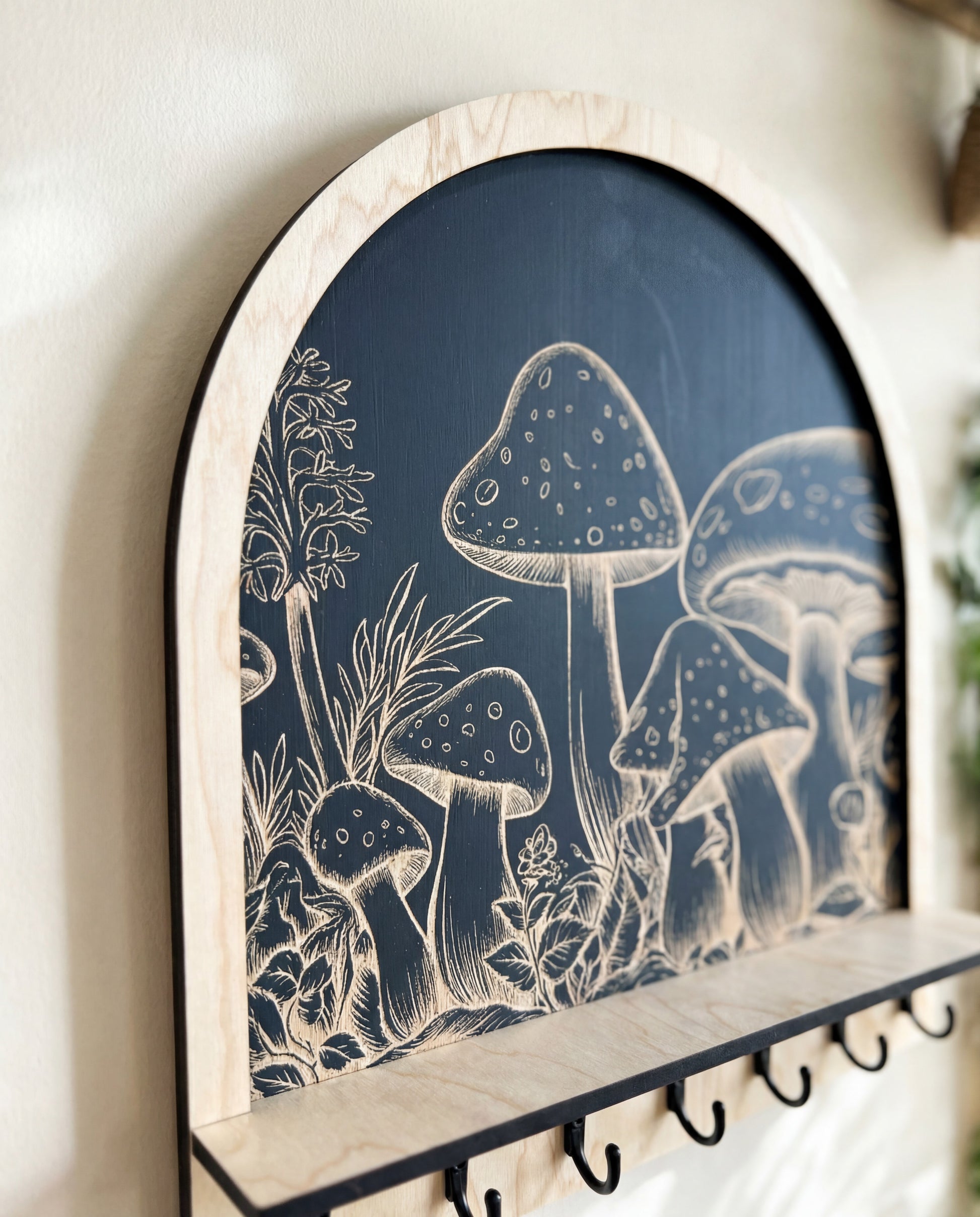 Up-close detail of an arched wooden wall key and jewelry holder featuring engraved mushroom and botanical artwork on a dark background, with a small shelf and black metal hooks.