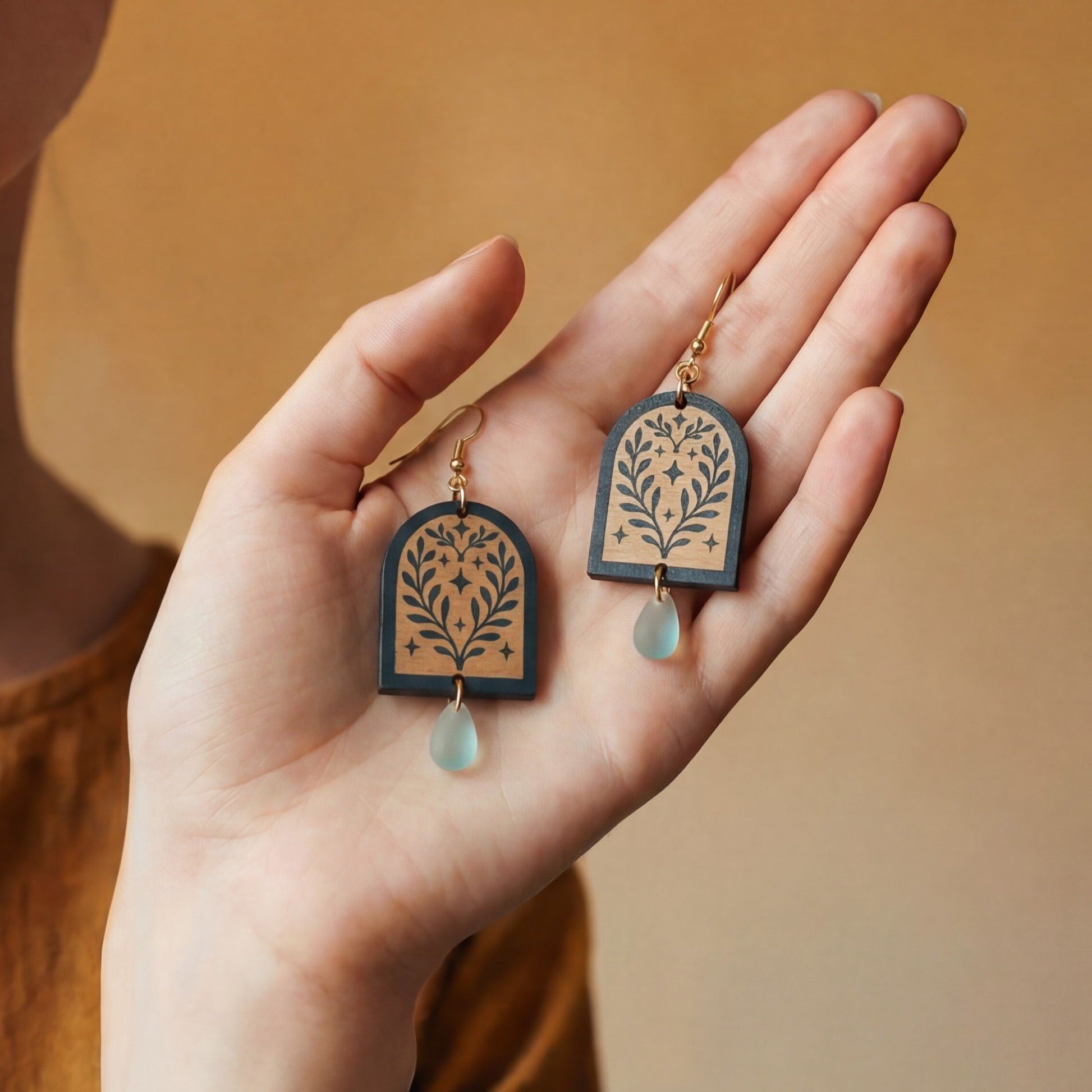 Hand holding a pair of green painted wooden earrings with an arched floral design and teardrop-shaped bead at the bottom against a warm background.