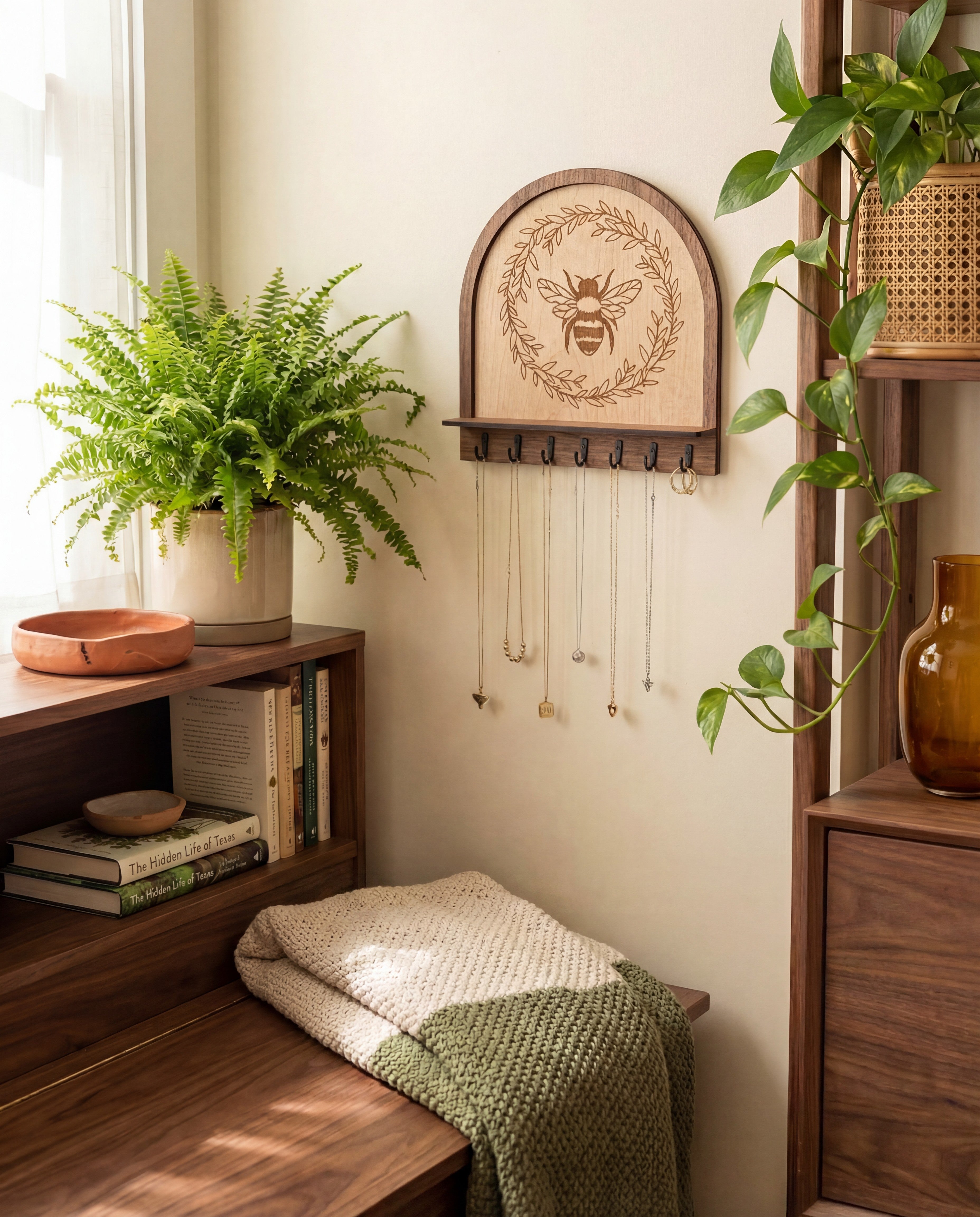 Maple wood bee wall organizer key holder with engraved honeybee wreath design and seven black hooks, styled on a neutral wall holding necklaces and jewelry in a cozy entryway nook with plants and wood shelving.