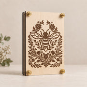 Laser-engraved wooden flower press featuring a detailed bee surrounded by floral vines and blossoms, finished with brass hardware on light maple wood.