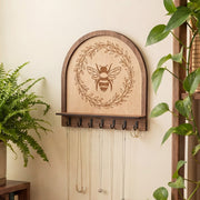 Maple wood bee wall organizer key holder with engraved honeybee wreath design, arched top and black metal hooks, shown holding necklaces and a key ring on a neutral wall with greenery nearby