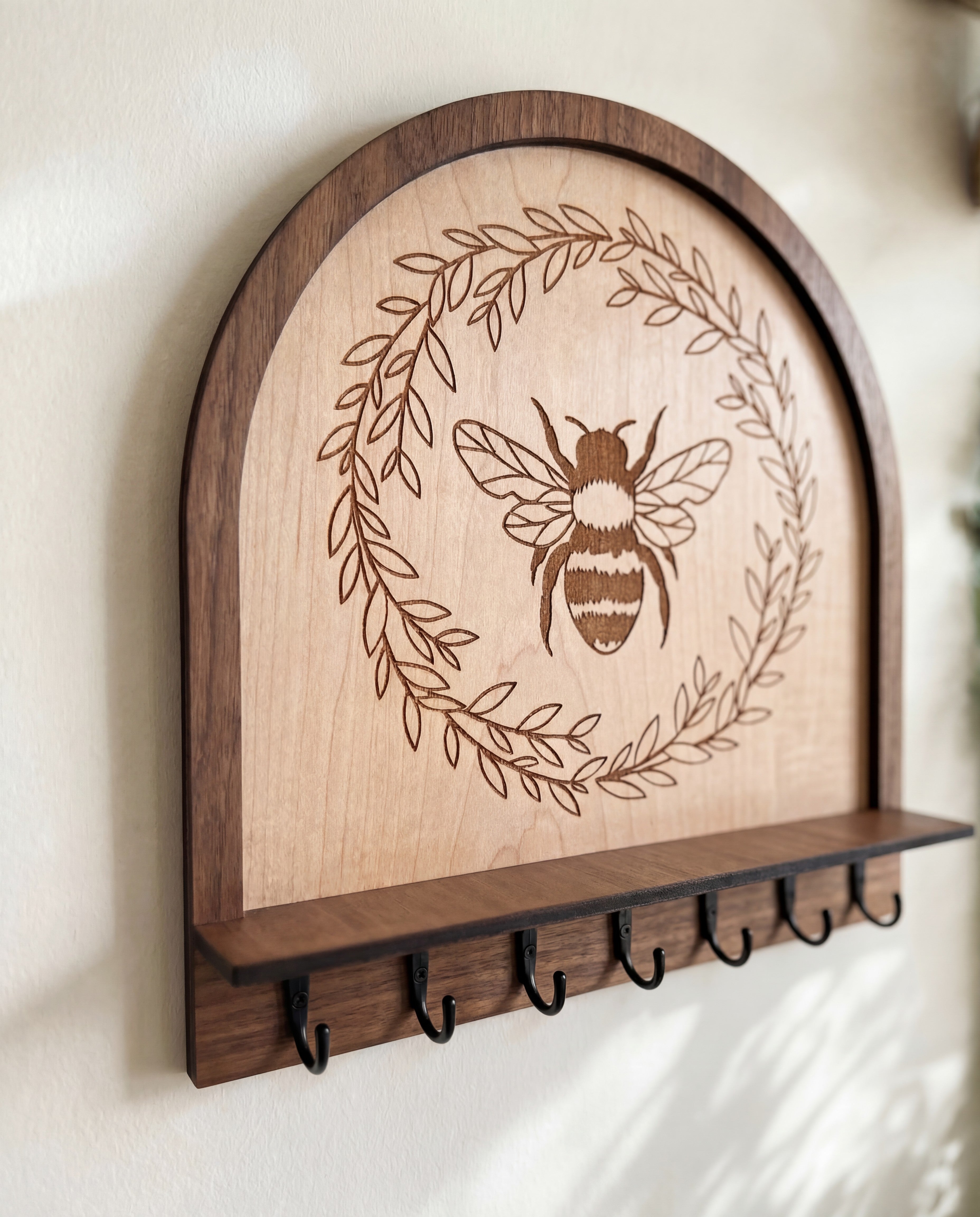 Maple wood bee wall organizer key holder with engraved honeybee wreath design, arched top and black metal hooks, on a neutral wall wall.