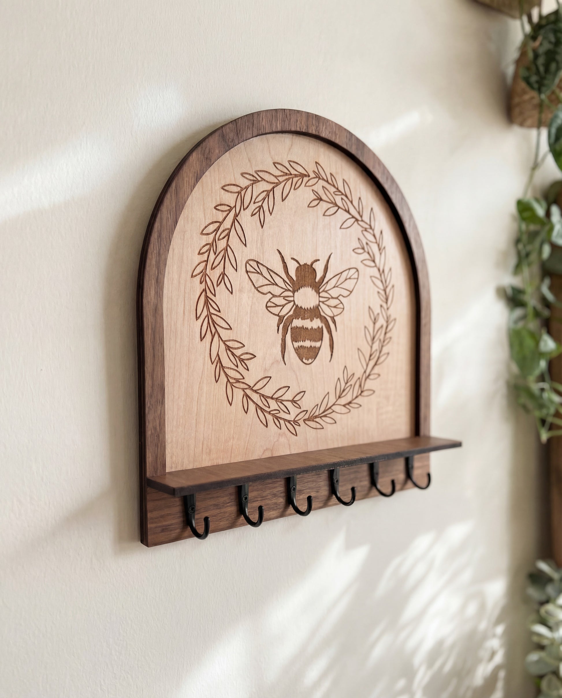 Close-up of maple wood bee wall organizer key holder featuring an engraved honeybee wreath design, arched layered wood frame, and black metal hooks mounted on a neutral wall in soft natural light.