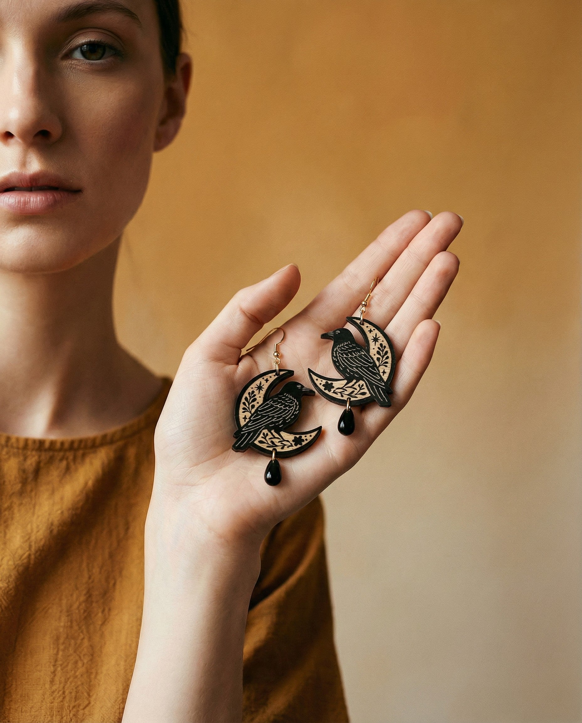 Person holding a pair of earrings with raven sitting on a moon design against a warm background