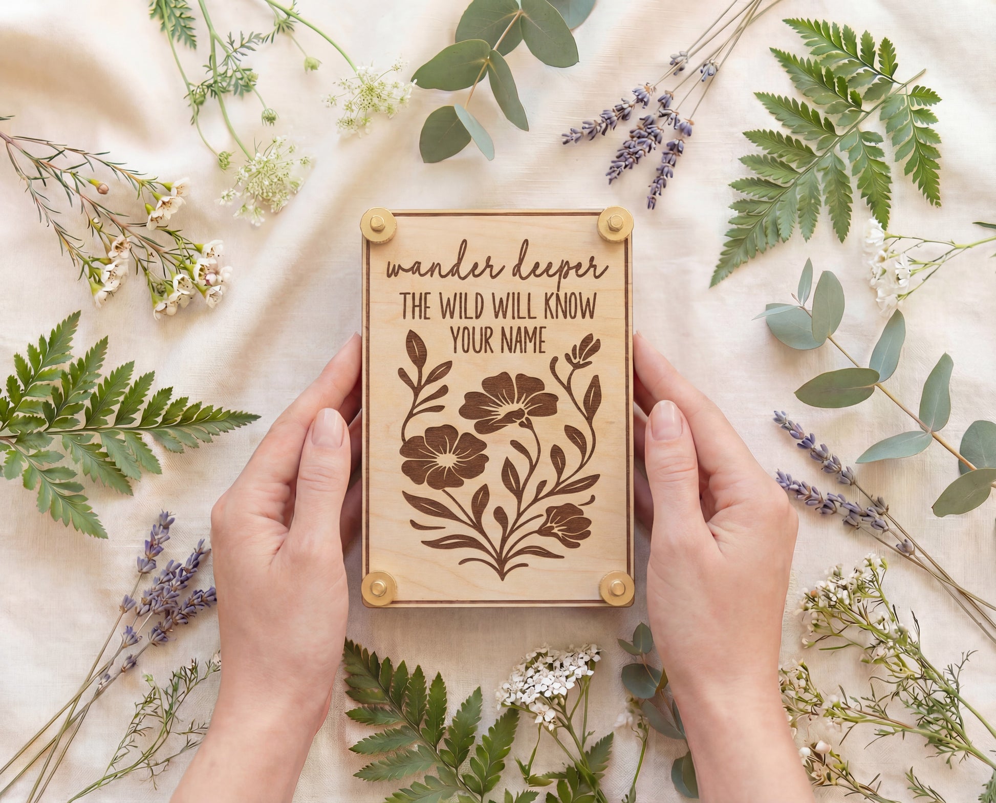 Handmade 4x6 maple flower press with brass hardware and “Wander deeper, the wild will know your name” engraved floral design, held in hands on cream linen surrounded by ferns, eucalyptus, lavender sprigs, and wildflowers.