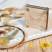Laser engraved wooden flower press with a fern and floral design, shown with metal bolts and styled with dried flowers on a rustic wood surface.