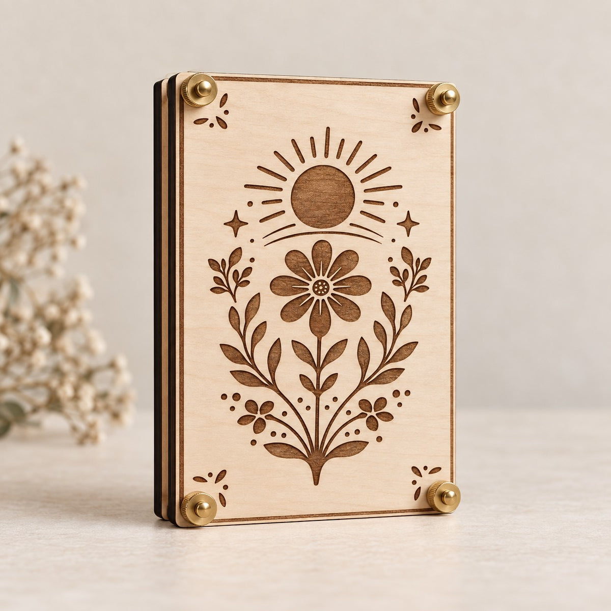 A rectangular wooden flower press with a laser-engraved floral and sun design, standing upright on a light surface. The natural wood has brass-colored corner hardware, and a softly blurred dried flower arrangement appears on the left side.
