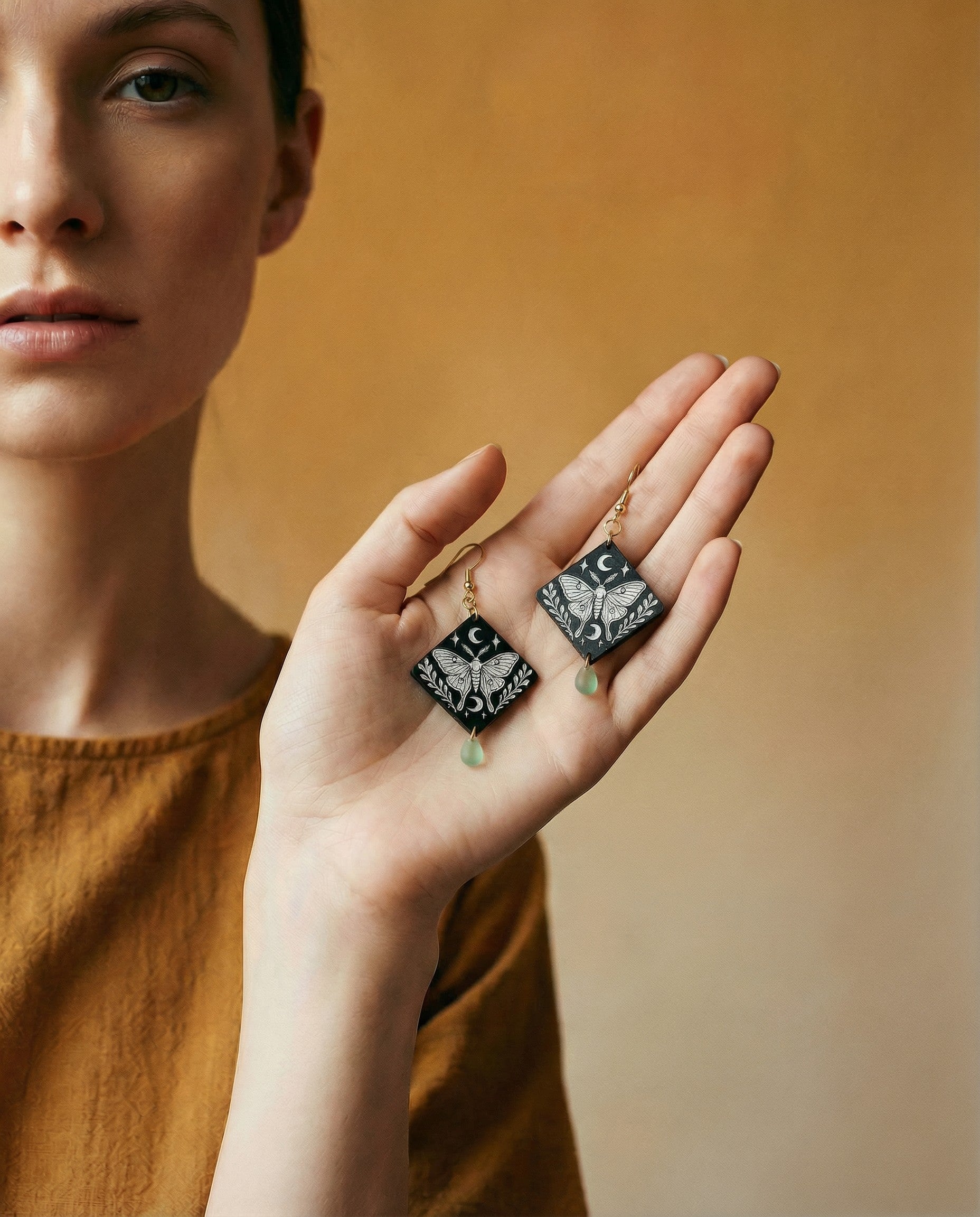 Person holding engraved moth diamond wood dangle earrings with crescent moon details, mint Czech glass teardrop beads, and 14k gold hooks.