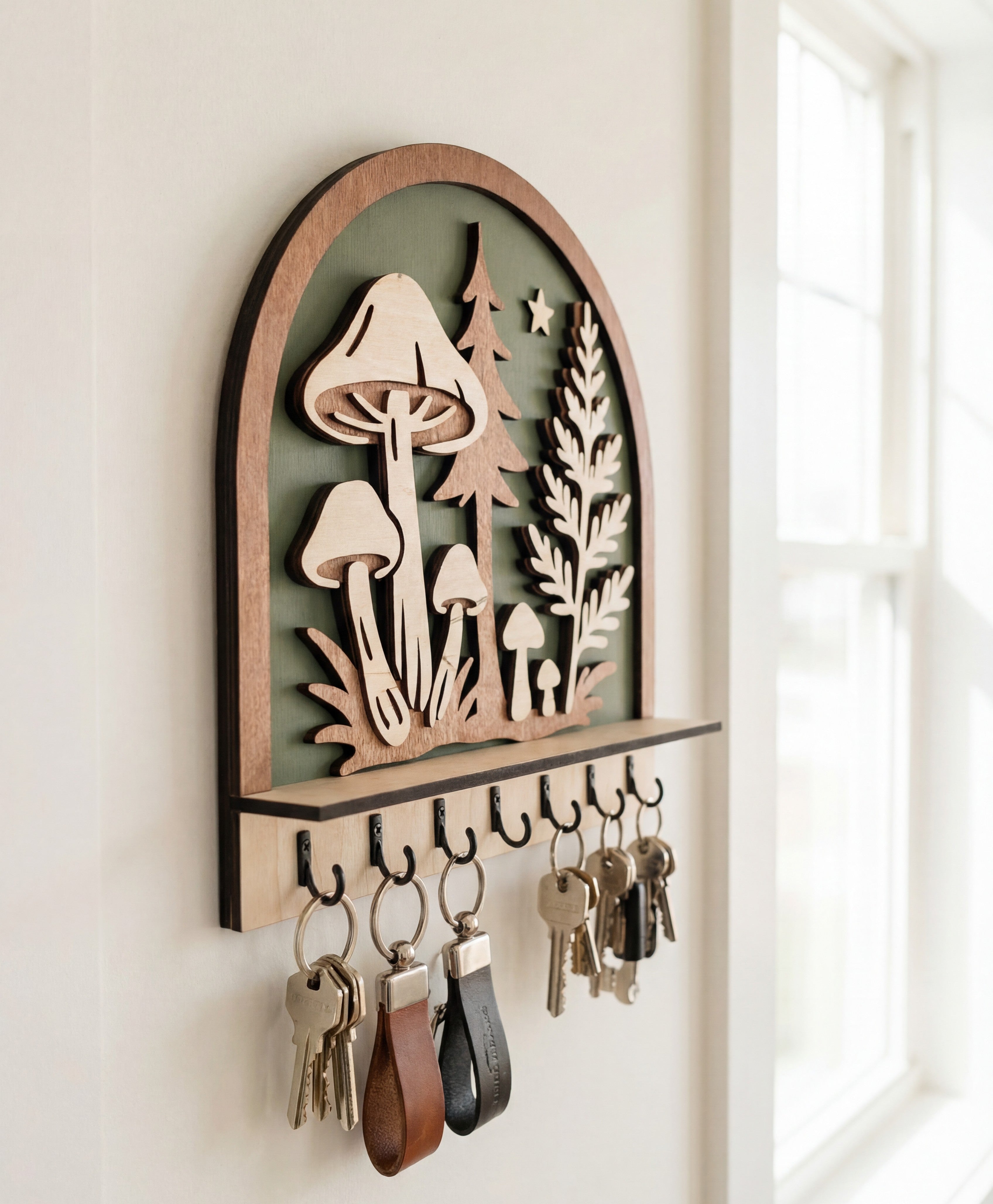 Wooden arched wall key holder with mushroom and forest cutout design, featuring multiple hooks holding keyrings, mounted beside a bright window.
