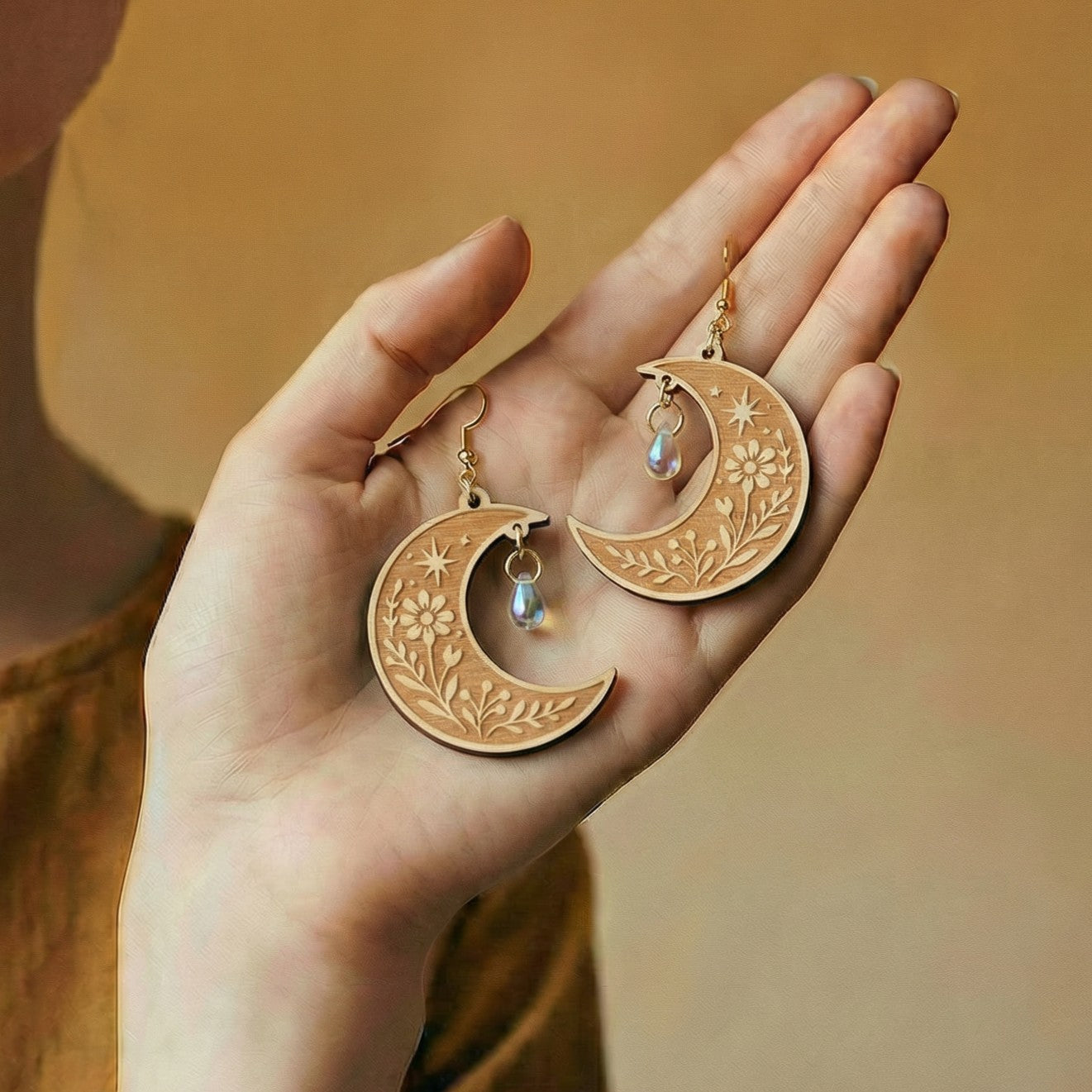 Hand holding gold moon-shaped earrings with floral designs and small stones against a warm background