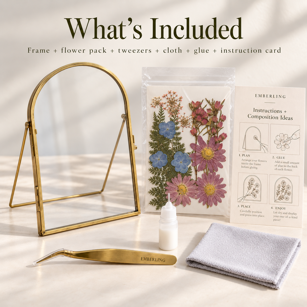 Embering floral arrangement kit components displayed on a light marble surface under warm sunlight, including a gold metal frame, a small mirror, tweezers, a folded gray cloth, a white glue bottle, a packet of pressed flowers, and an instruction card. Text at the top reads 'What's Included'.