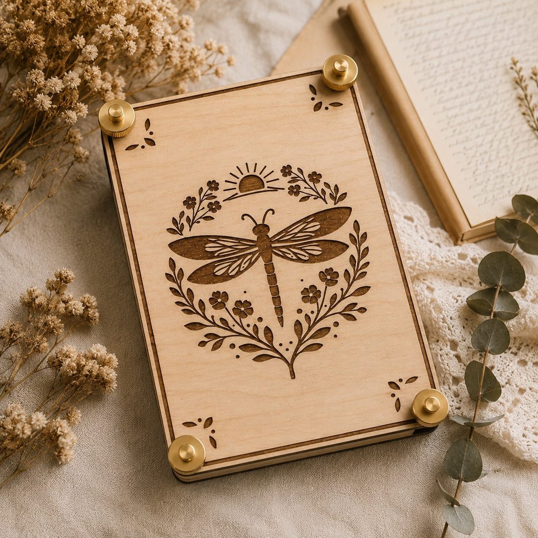 Handmade wooden flower press with laser-engraved celestial dragonfly botanical design, brass hardware, styled with dried wildflowers and eucalyptus on linen