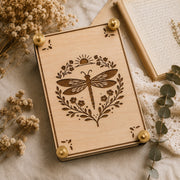 Handmade wooden flower press with laser-engraved celestial dragonfly botanical design, brass hardware, styled with dried wildflowers and eucalyptus on linen