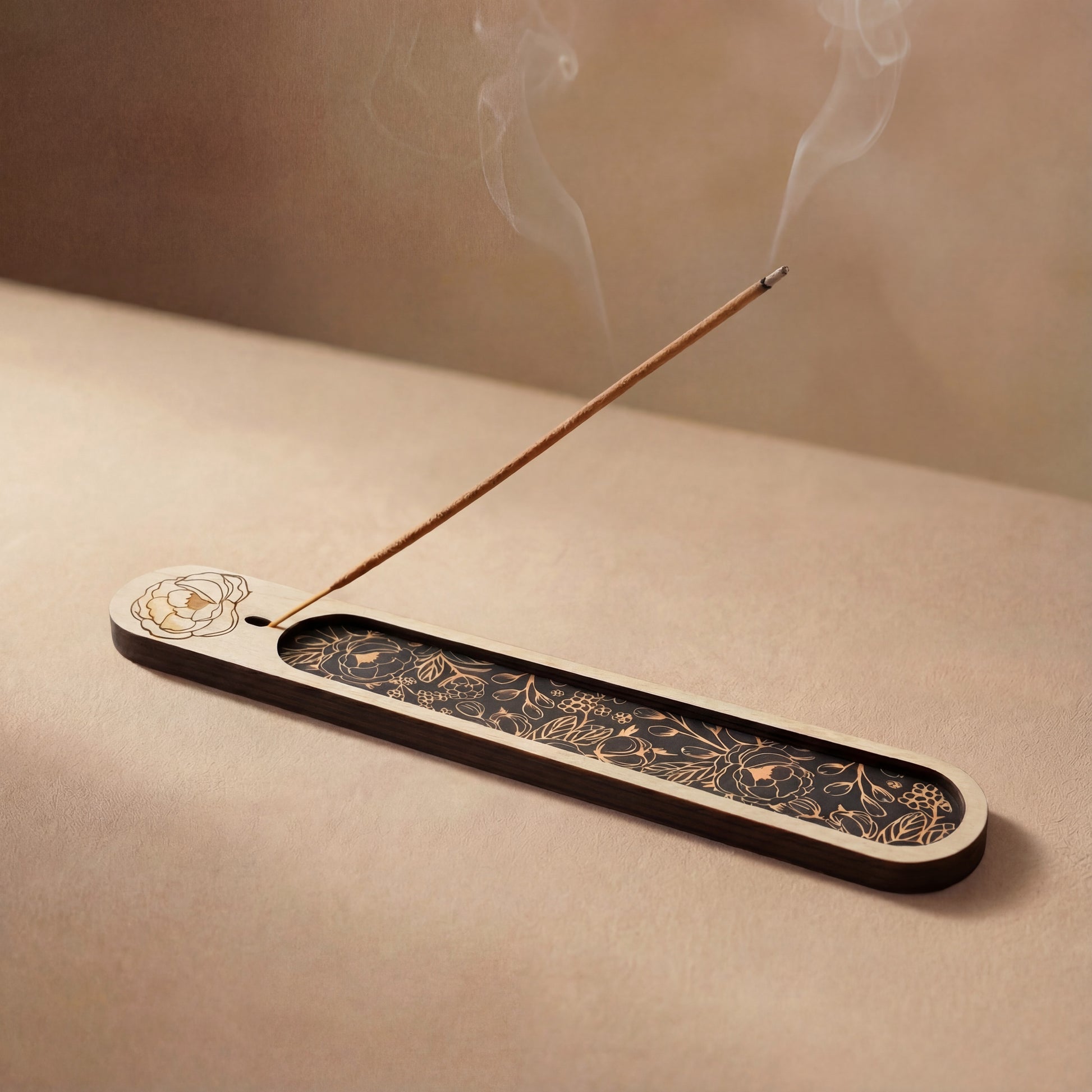 Wooden incense holder with a floral engraving and black botanical inlay, holding a lit incense stick with smoke rising on a neutral background.