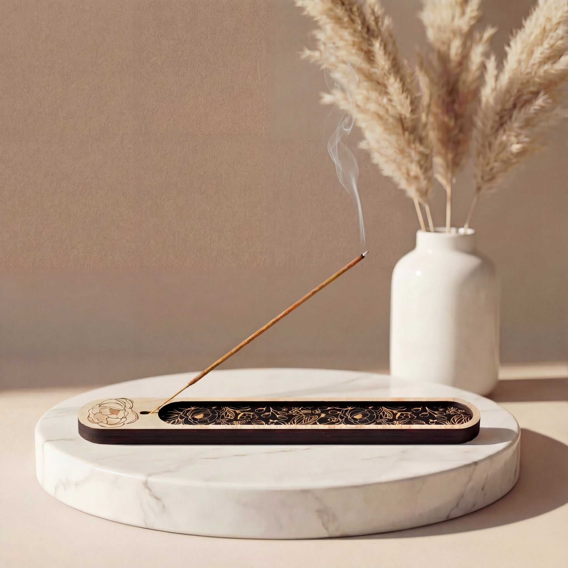 Wooden incense holder with a black botanical inlay holding a lit incense stick, styled on a round marble tray with a white vase of dried pampas grass in the background.