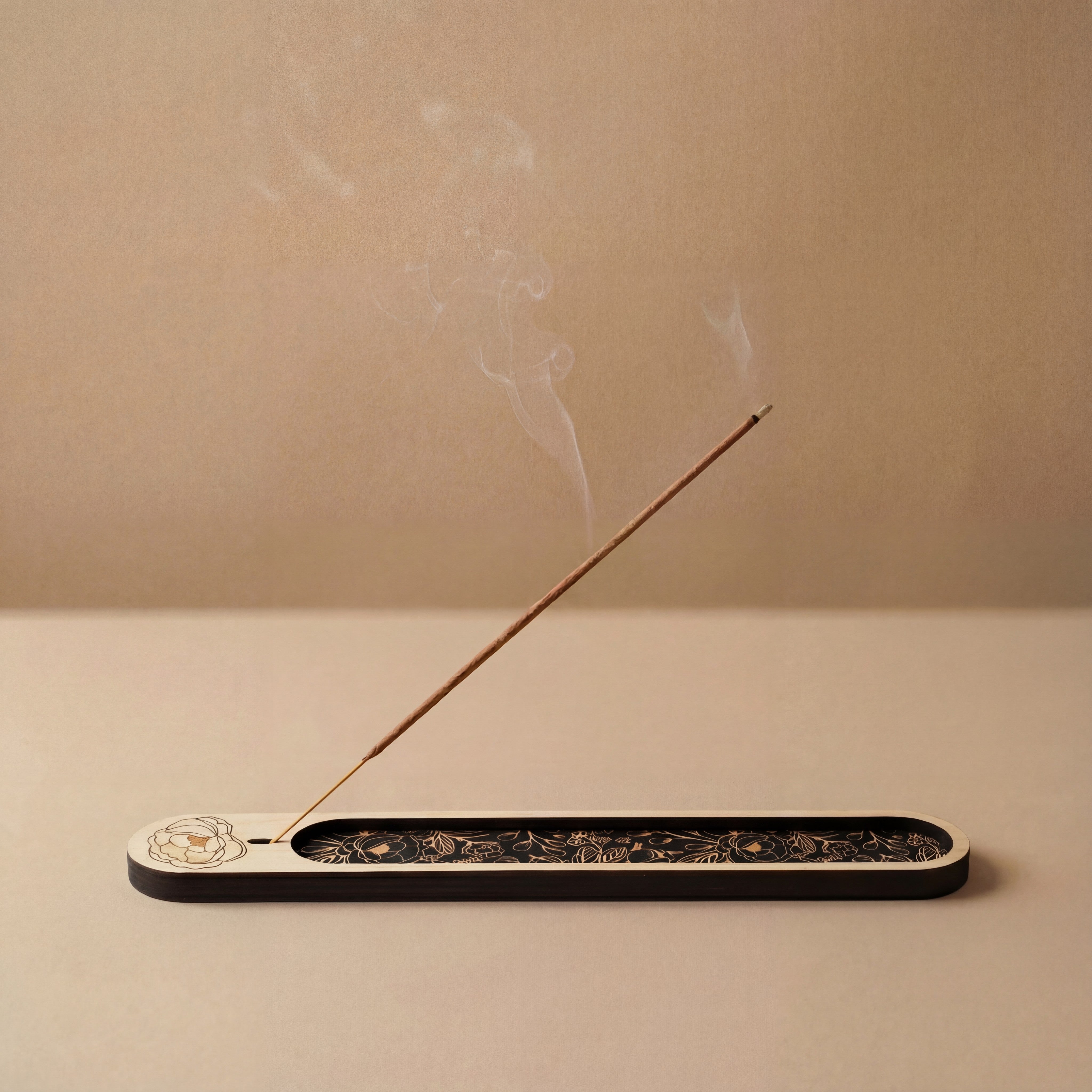 Minimalist wooden incense holder with a black floral inlay holding a lit incense stick, with soft smoke rising against a neutral background.