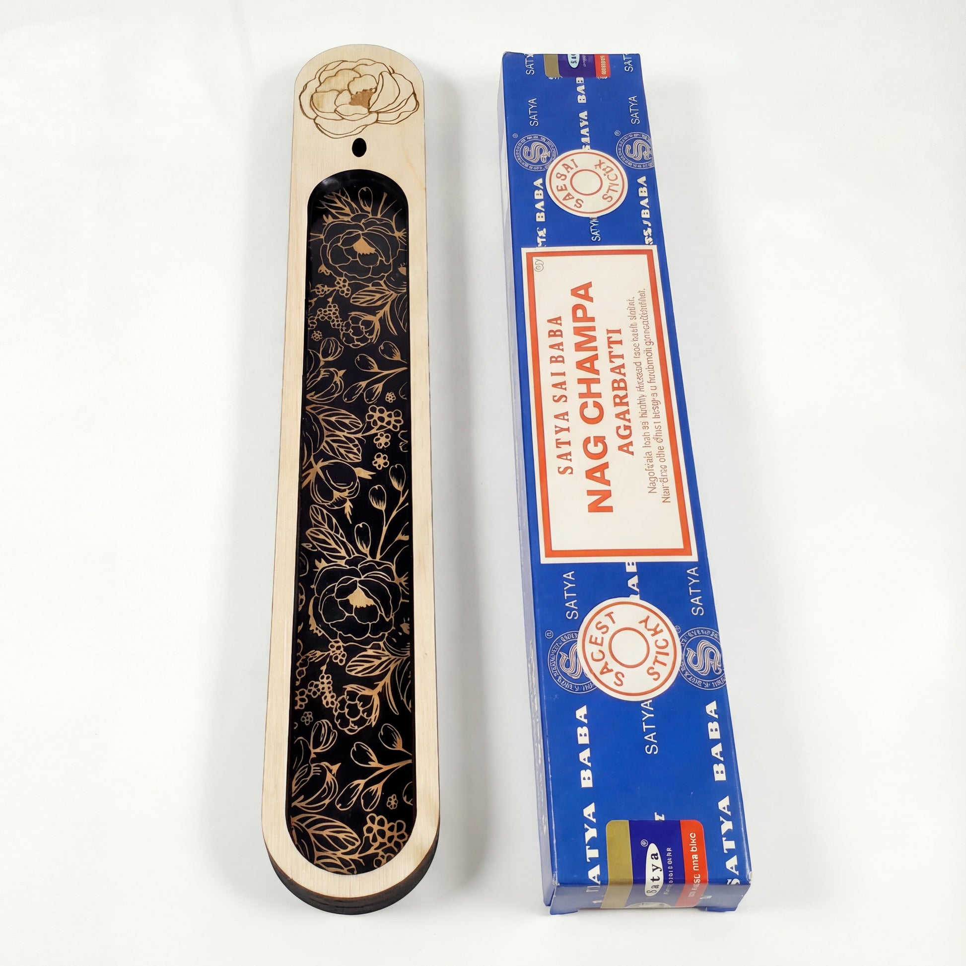 Top-down view of a wooden incense holder with a black floral inlay next to a blue Nag Champa incense box on a white background.