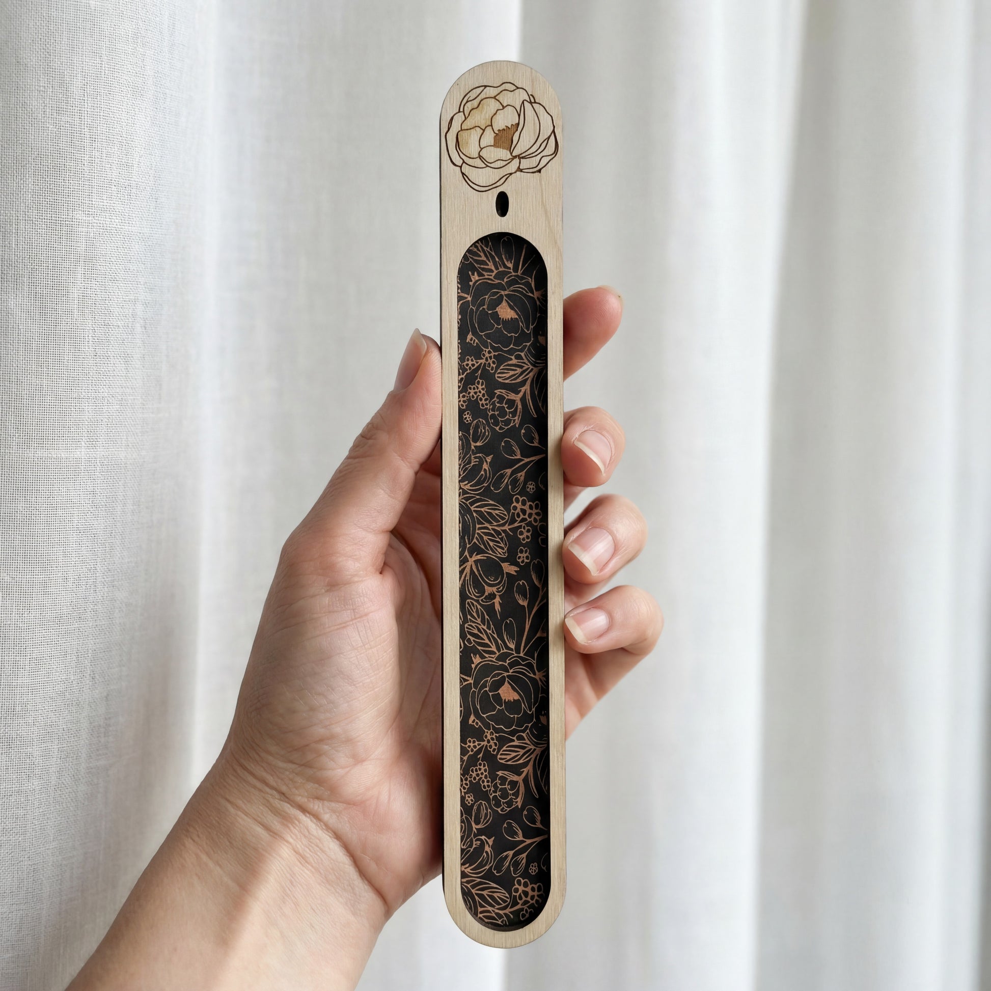 Hand holding a slim wooden bookmark with a black floral inlay and an engraved flower at the top, photographed against a white curtain.
