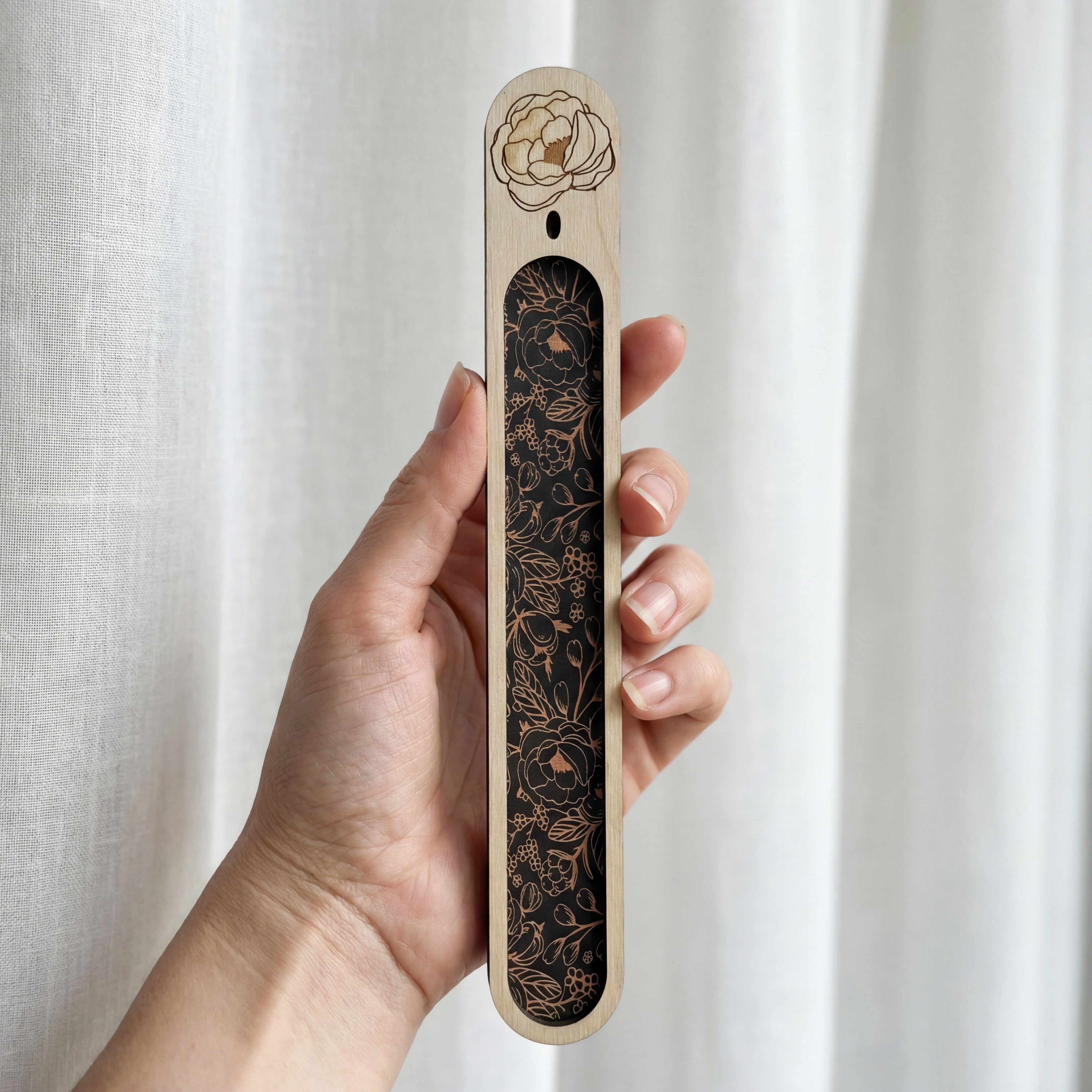 Hand holding a slim wooden bookmark with a black floral inlay and an engraved flower at the top, photographed against a white curtain.