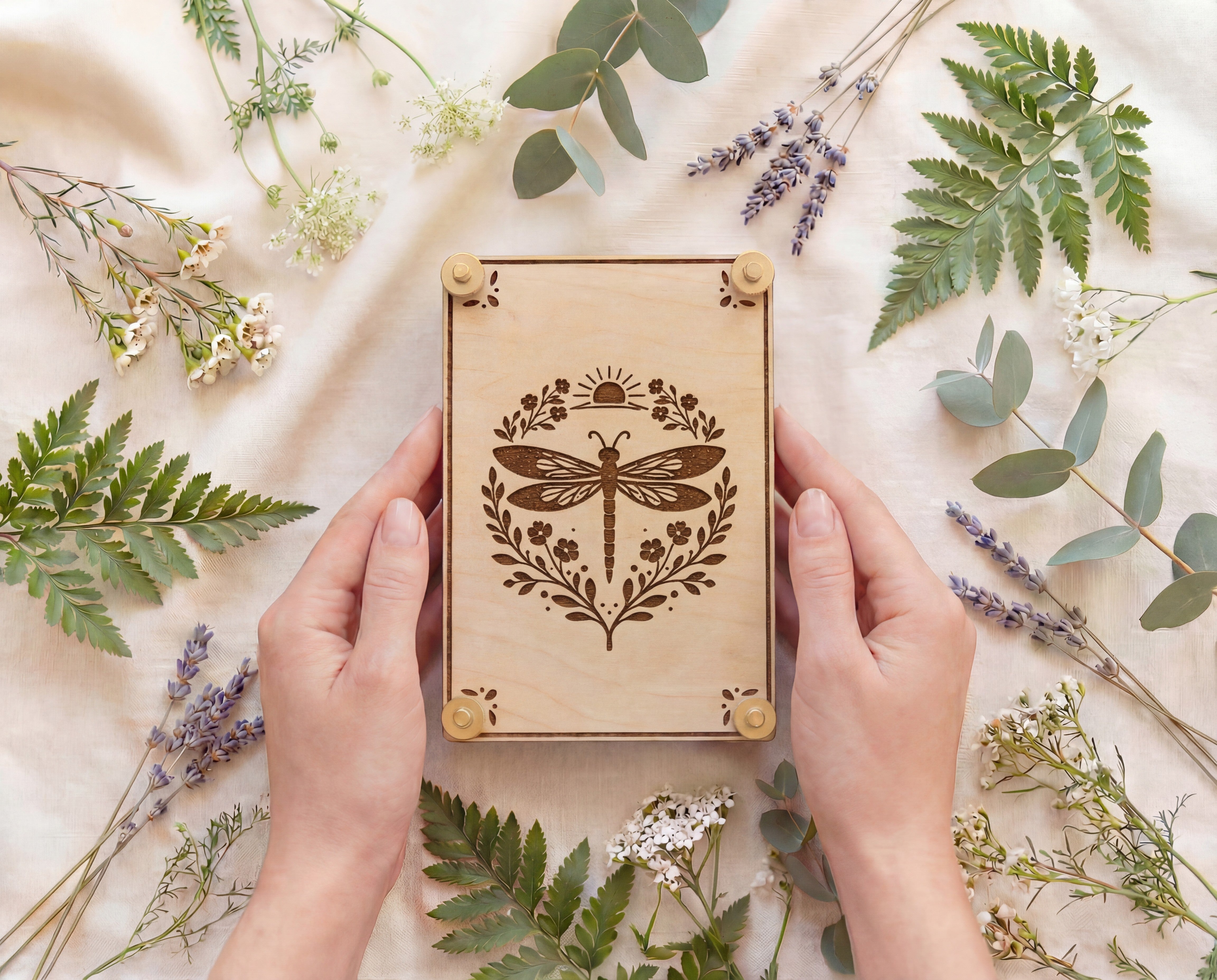 Hands holding a handmade 4x6 maple wooden flower press kit with engraved dragonfly botanical design and brass knurled thumb nuts, styled on a neutral linen backdrop surrounded by ferns, eucalyptus, lavender and wildflowers, a portable pressed flower kit for beginners for botanical crafts, scrapbooking, nature journaling and pressed flower art.
