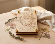 Handmade 4x6 maple wood flower press with brass knurled thumb nuts, engraved woman line art with peonies and butterflies, styled with pressed flowers on a light wood table.