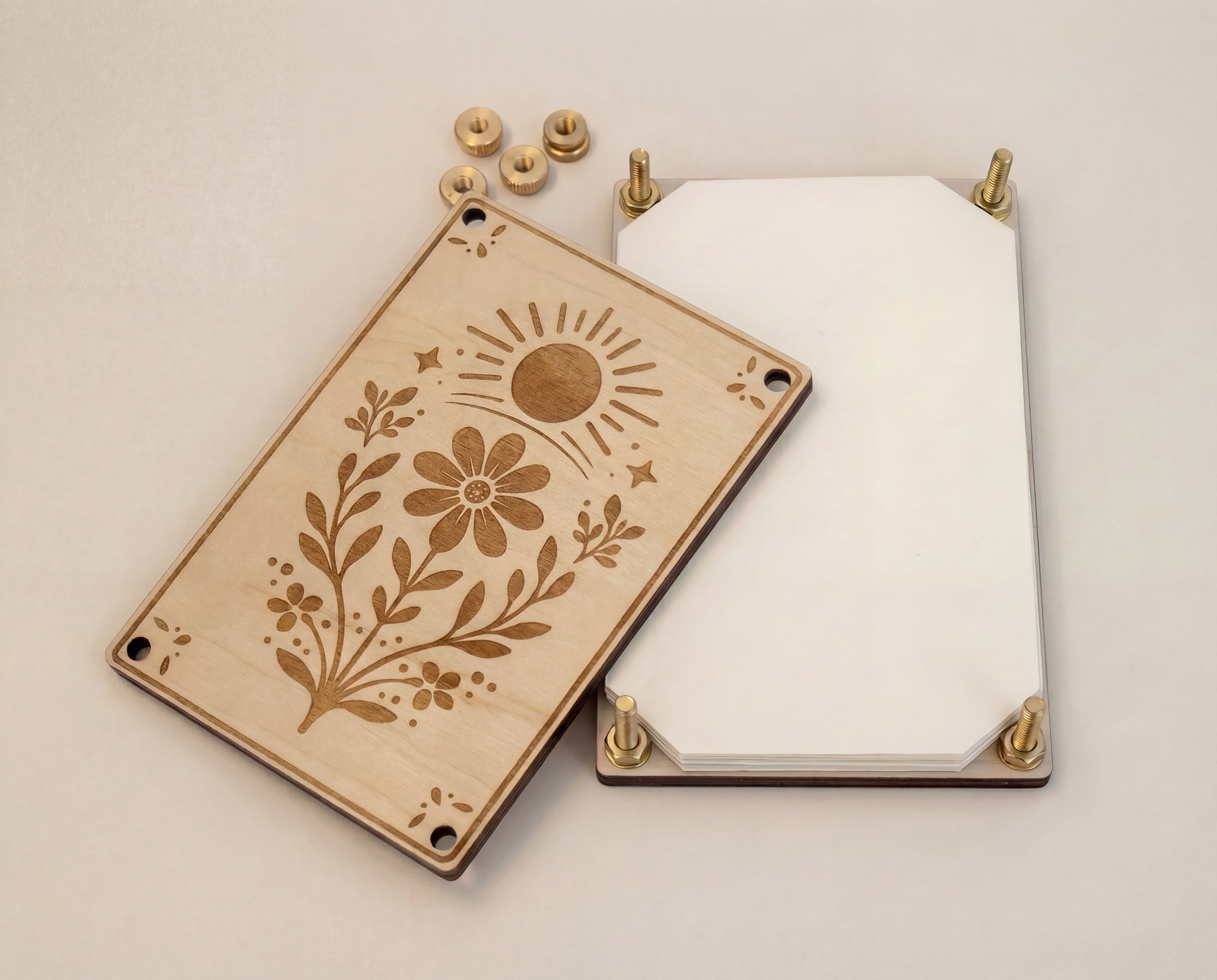 Handmade 4x6 maple wooden flower press kit with engraved sun and wildflower design, brass round bolts, washers, hex nuts and knurled thumb nuts, shown open with stacked white blotting paper and cardboard inserts for pressing wildflowers and leaves for pressed flower art, nature journaling and botanical crafts.