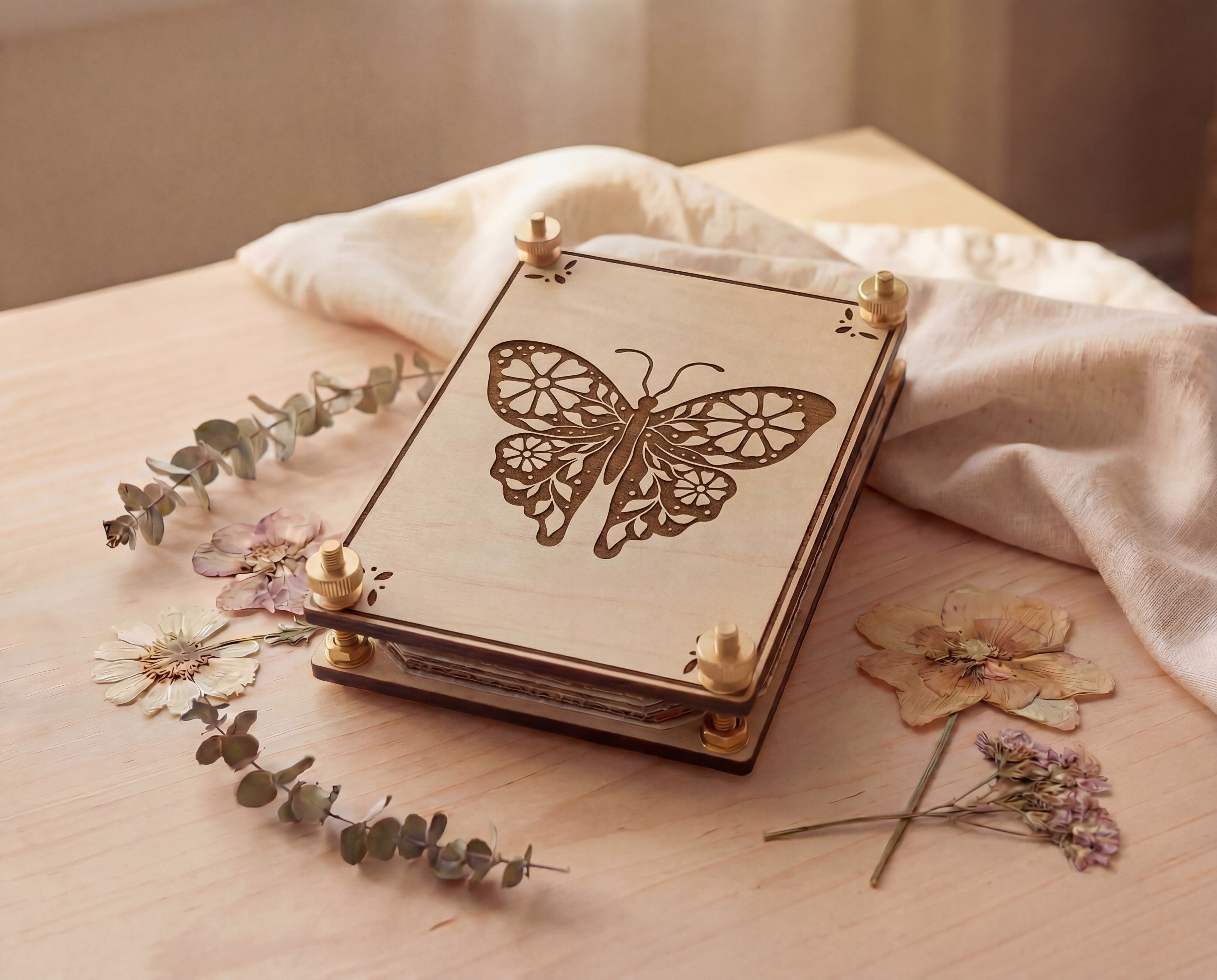 Handmade wooden flower press with laser-cut butterfly design and brass screw hardware, photographed at an angle on a light wood table with eucalyptus sprigs and pressed wildflowers; reusable eco-friendly botanical flower pressing kit for drying flowers for crafts, journaling, resin art, and a nature-inspired gift for plant lovers.