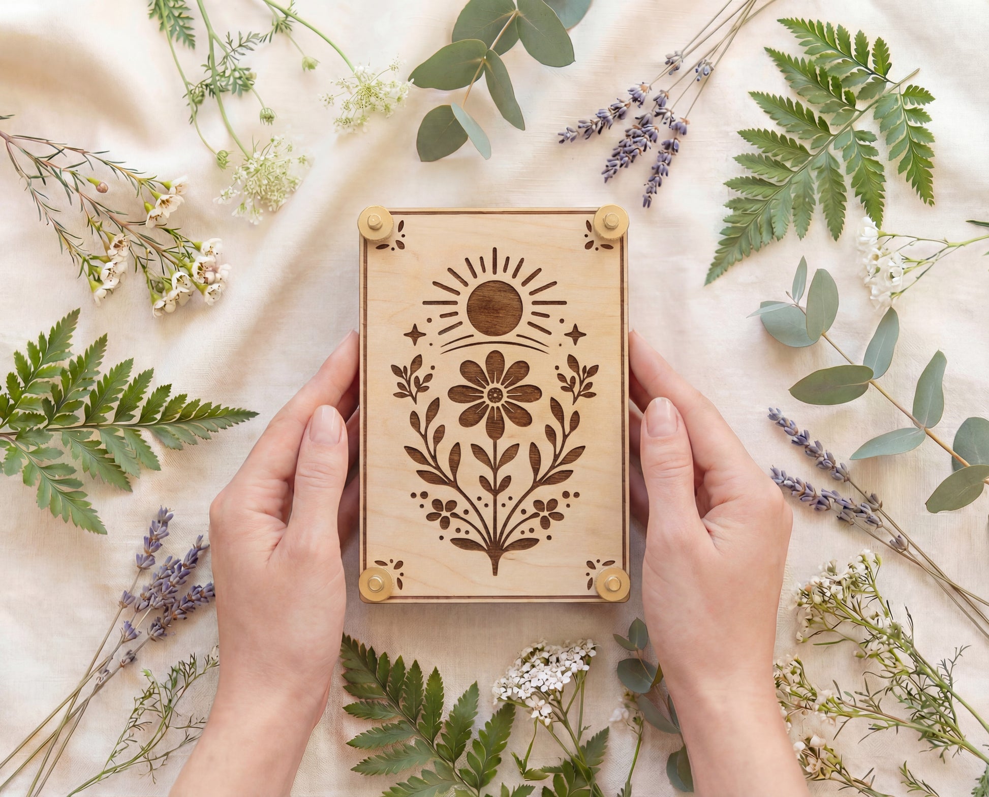 Hands holding a handmade 4x6 maple wooden flower press kit with engraved sun and wildflower botanical design and brass knurled thumb nuts, surrounded by fresh greenery, fern fronds, eucalyptus and lavender on a neutral linen backdrop, a portable pressed flower kit for beginners to press wildflowers and leaves for botanical crafts, scrapbooking, nature journaling and pressed flower art.