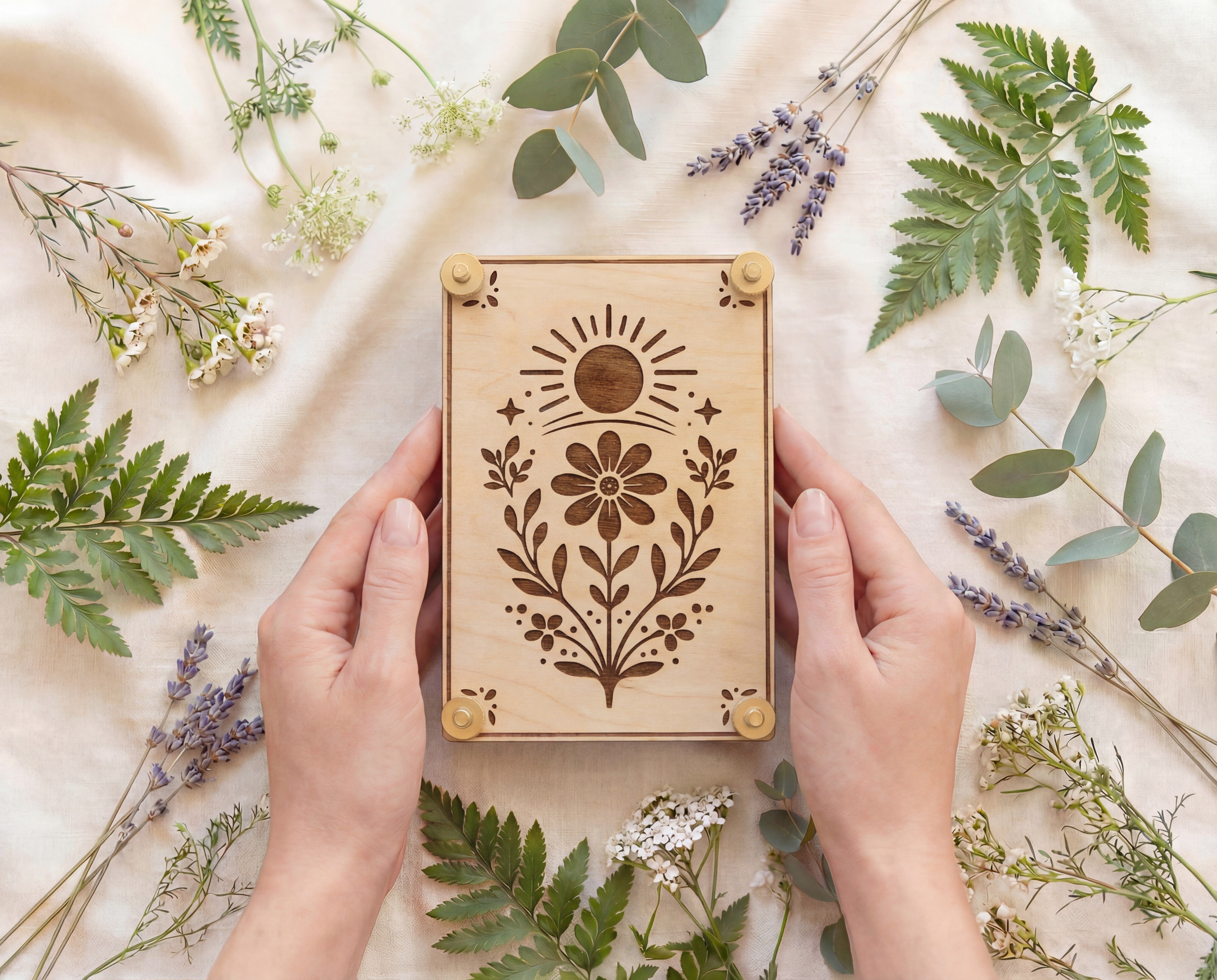 Hands holding a handmade 4x6 maple wooden flower press kit with engraved sun and wildflower botanical design and brass knurled thumb nuts, surrounded by fresh greenery, fern fronds, eucalyptus and lavender on a neutral linen backdrop, a portable pressed flower kit for beginners to press wildflowers and leaves for botanical crafts, scrapbooking, nature journaling and pressed flower art.