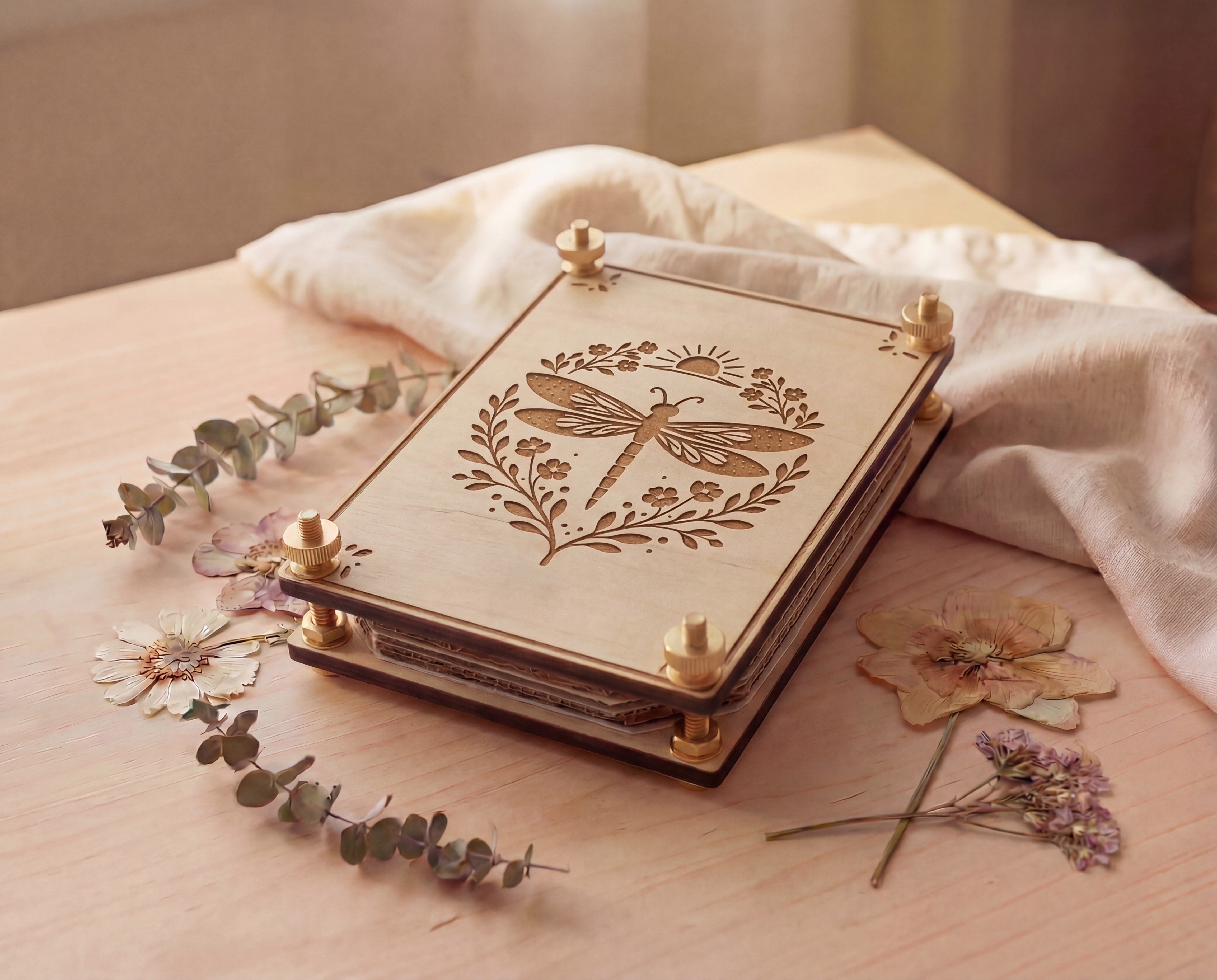 Angled view of a handmade 4x6 maple wooden flower press kit with an engraved dragonfly botanical design and brass knurled thumb nuts, assembled with brass round bolts, washers and hex nuts, styled on a light wood table with dried pressed flowers and eucalyptus beside soft linen fabric, a portable pressed flower kit for beginners for pressing wildflowers and leaves for botanical crafts, scrapbooking, nature journaling and pressed flower art.