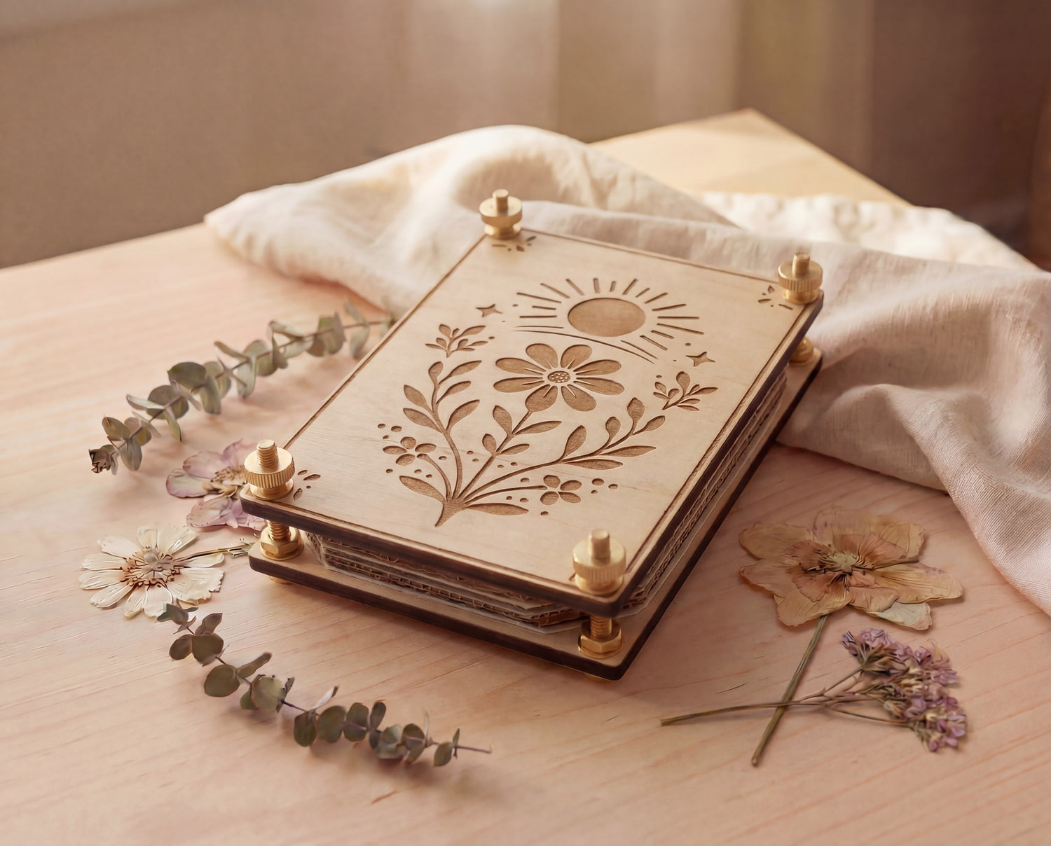 Angled view of a handmade 4x6 maple wooden flower press kit with engraved sun and wildflower botanical design, assembled with brass round bolts, washers, hex nuts and knurled thumb nuts, sitting on a light wood table beside dried pressed flowers and eucalyptus with soft linen fabric, a portable pressed flower kit for beginners for pressing wildflowers and leaves for botanical crafts, scrapbooking, nature journaling and pressed flower art.