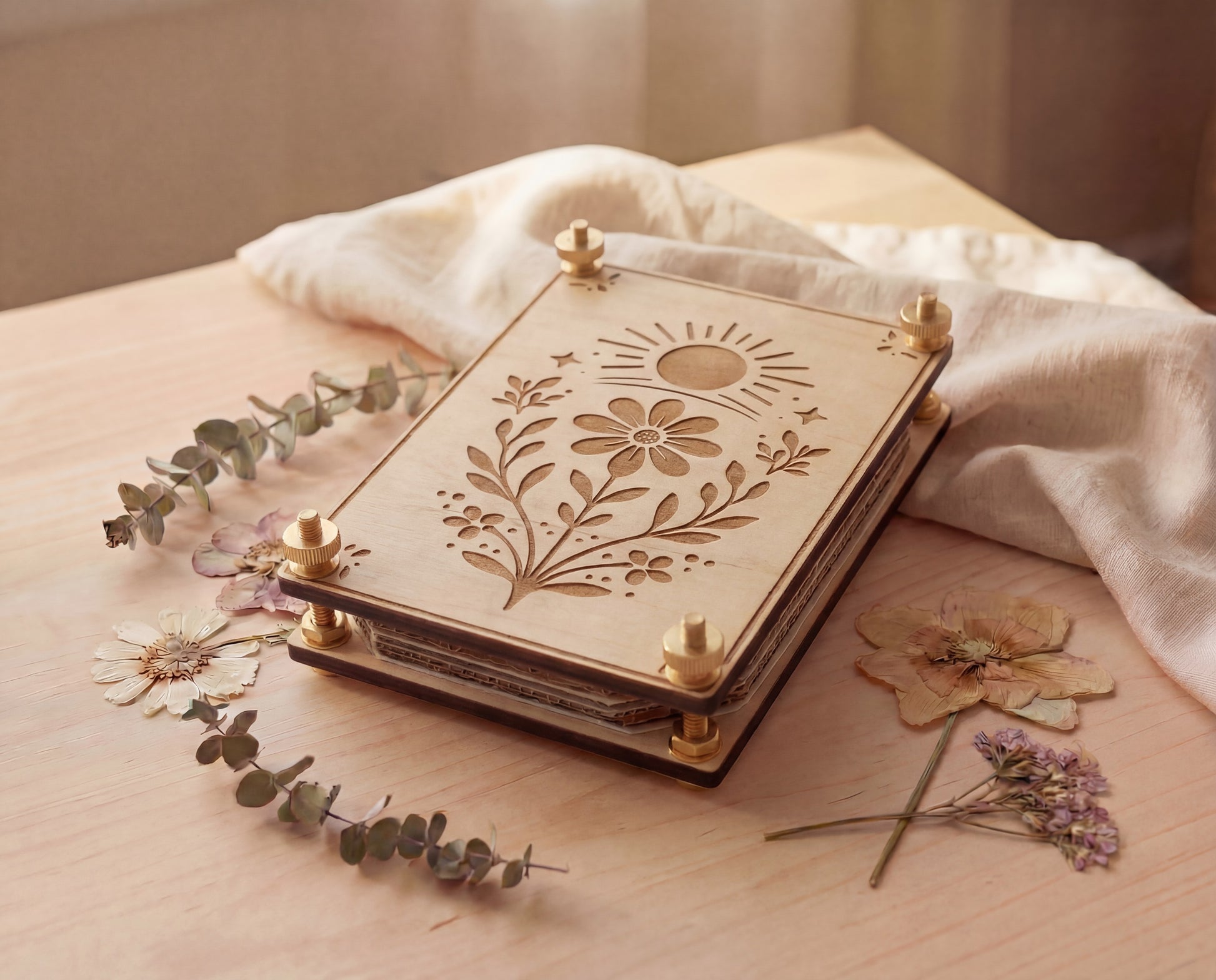 Angled view of a handmade 4x6 maple wooden flower press kit with engraved sun and wildflower botanical design, assembled with brass round bolts, washers, hex nuts and knurled thumb nuts, sitting on a light wood table beside dried pressed flowers and eucalyptus with soft linen fabric, a portable pressed flower kit for beginners for pressing wildflowers and leaves for botanical crafts, scrapbooking, nature journaling and pressed flower art.