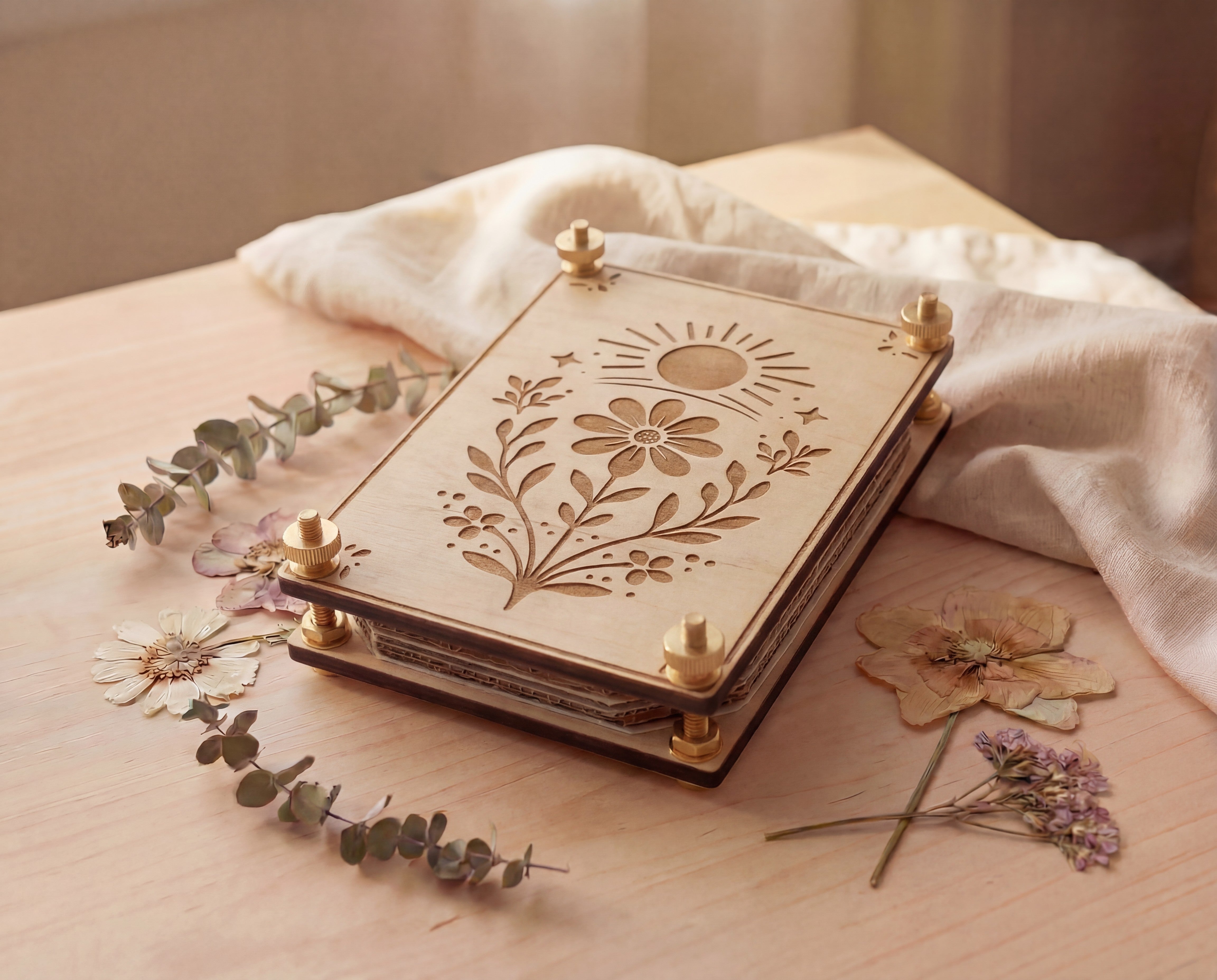 Angled view of a handmade 4x6 maple wooden flower press kit with engraved sun and wildflower botanical design, assembled with brass round bolts, washers, hex nuts and knurled thumb nuts, sitting on a light wood table beside dried pressed flowers and eucalyptus with soft linen fabric, a portable pressed flower kit for beginners for pressing wildflowers and leaves for botanical crafts, scrapbooking, nature journaling and pressed flower art.