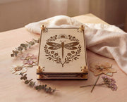 Front view of a handmade 4x6 maple wooden flower press kit with engraved dragonfly botanical design and brass knurled thumb nuts, stacked with blotting paper and cardboard inserts, styled on a light wood table with dried pressed flowers and eucalyptus beside soft linen fabric, a portable pressed flower kit for beginners for botanical crafts, scrapbooking, nature journaling and pressed flower art.