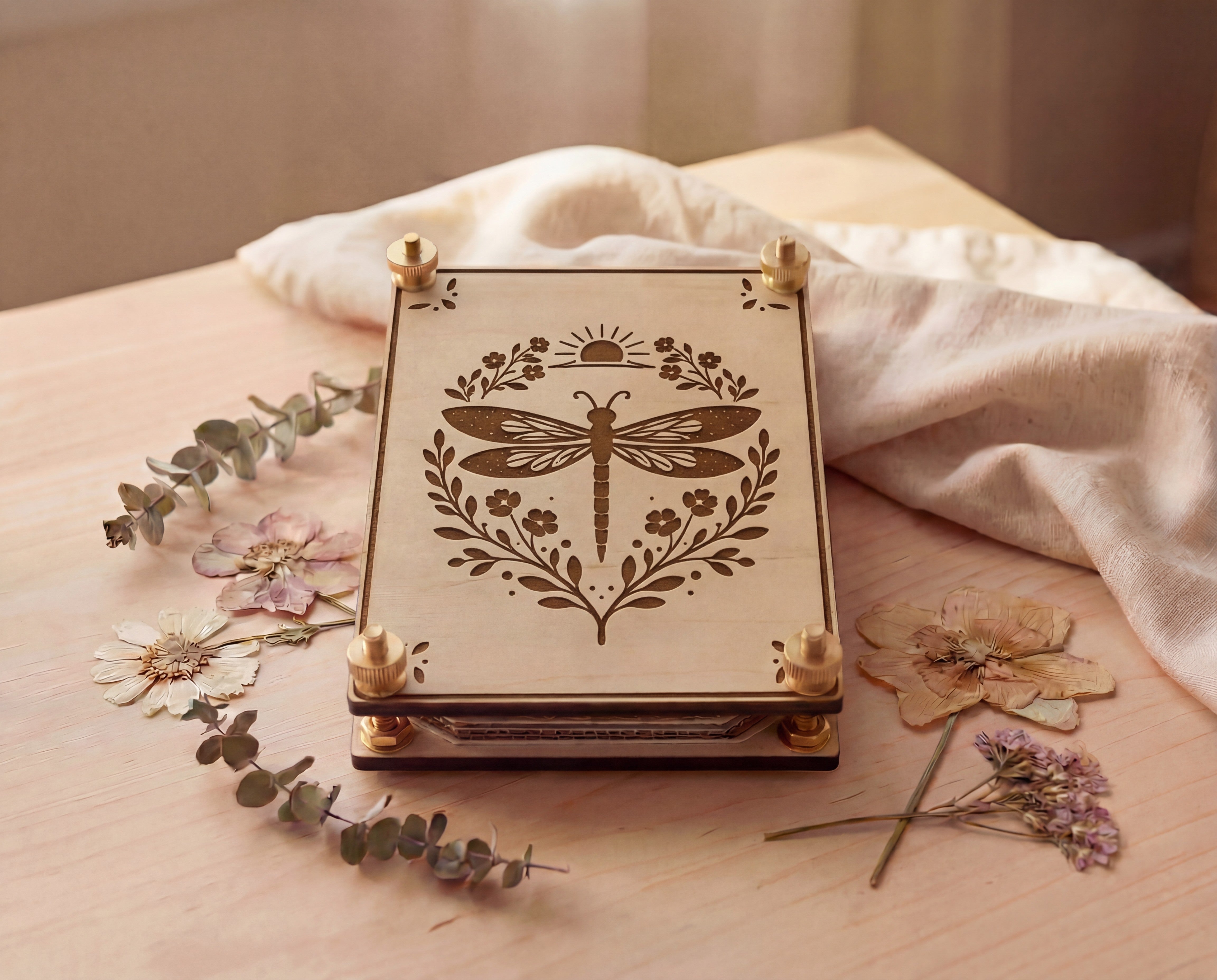 Front view of a handmade 4x6 maple wooden flower press kit with engraved dragonfly botanical design and brass knurled thumb nuts, stacked with blotting paper and cardboard inserts, styled on a light wood table with dried pressed flowers and eucalyptus beside soft linen fabric, a portable pressed flower kit for beginners for botanical crafts, scrapbooking, nature journaling and pressed flower art.