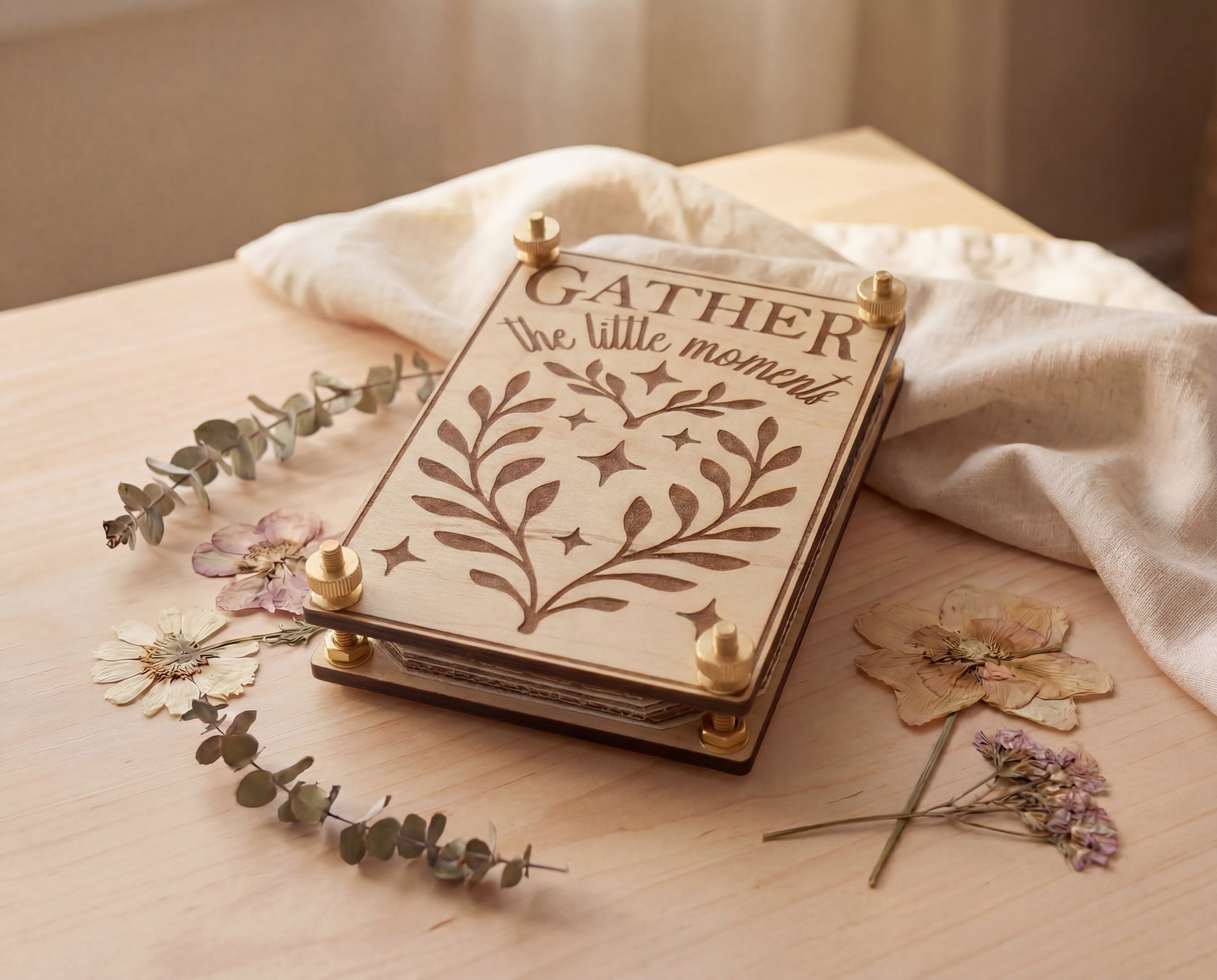 Handmade 4x6 maple flower press with brass hardware and original “Gather the little moments” engraved design, styled on a light wood table with linen cloth, eucalyptus sprigs, and pressed flowers.
