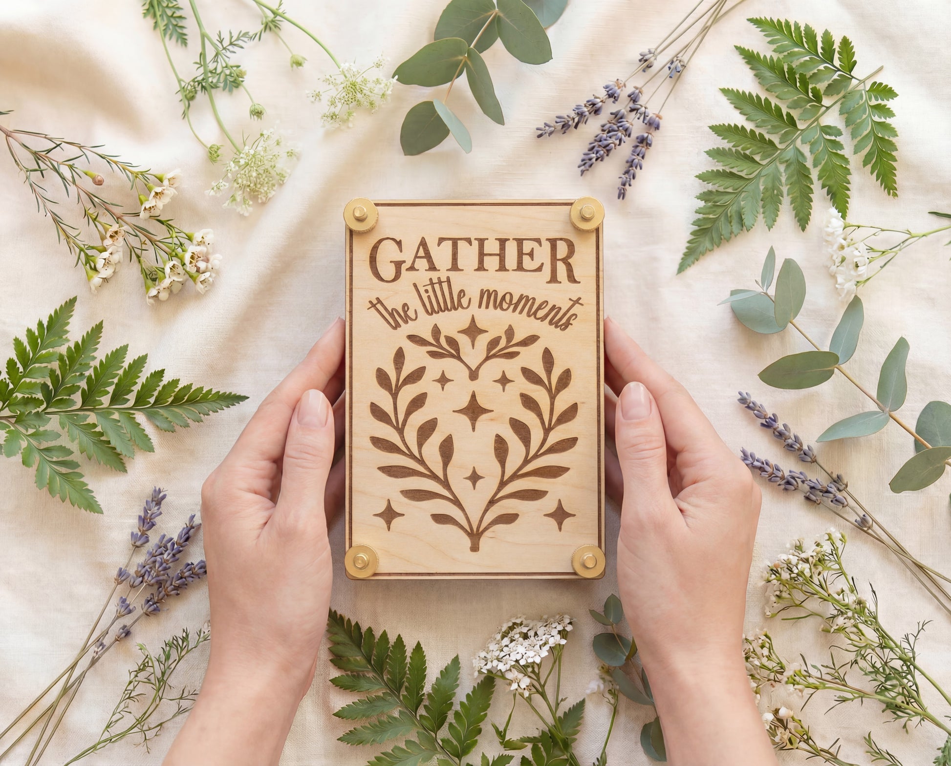 Handmade 4x6 maple flower press with brass hardware and original “Gather the little moments” engraved design, held in hands on cream linen surrounded by ferns, eucalyptus, lavender, and wildflowers.