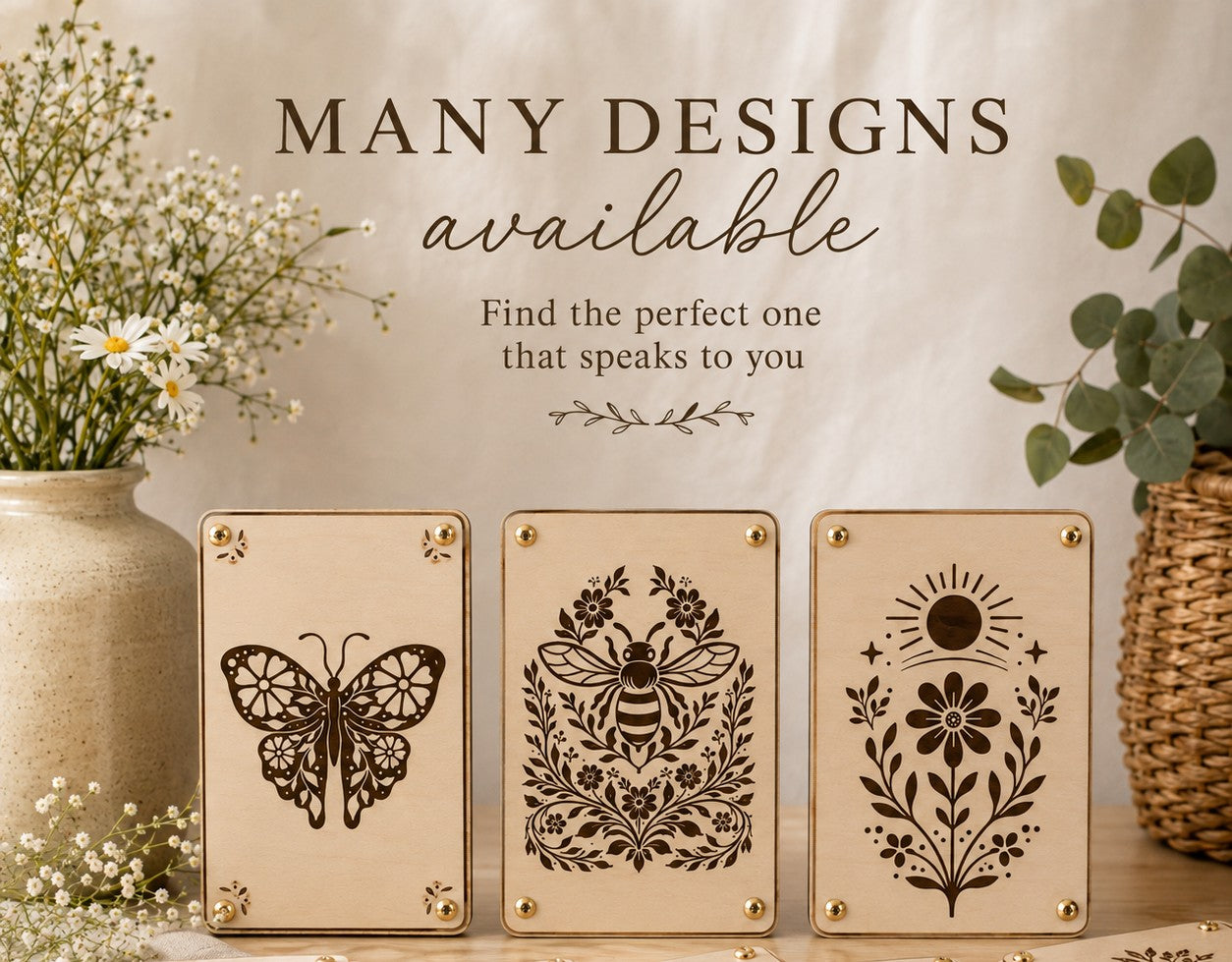 Three handmade wooden flower presses with laser-engraved designs displayed upright — intricate butterfly with floral wings, honeybee surrounded by folk art botanicals, and a celestial sun with wildflower bloom — styled with daisies, baby's breath, and eucalyptus.