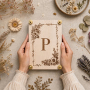 Close-up of hands holding a handmade wooden flower press featuring a laser-engraved monogram letter P with an intricate botanical floral corner border and brass hardware, styled on a linen surface with dried daisies, lavender, baby's breath, and eucalyptus.