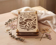 Handmade 4x6 maple flower press with brass hardware and “Wander deeper, the wild will know your name” engraved floral design, styled on a light wood table with linen cloth, eucalyptus sprigs, and pressed flowers.
