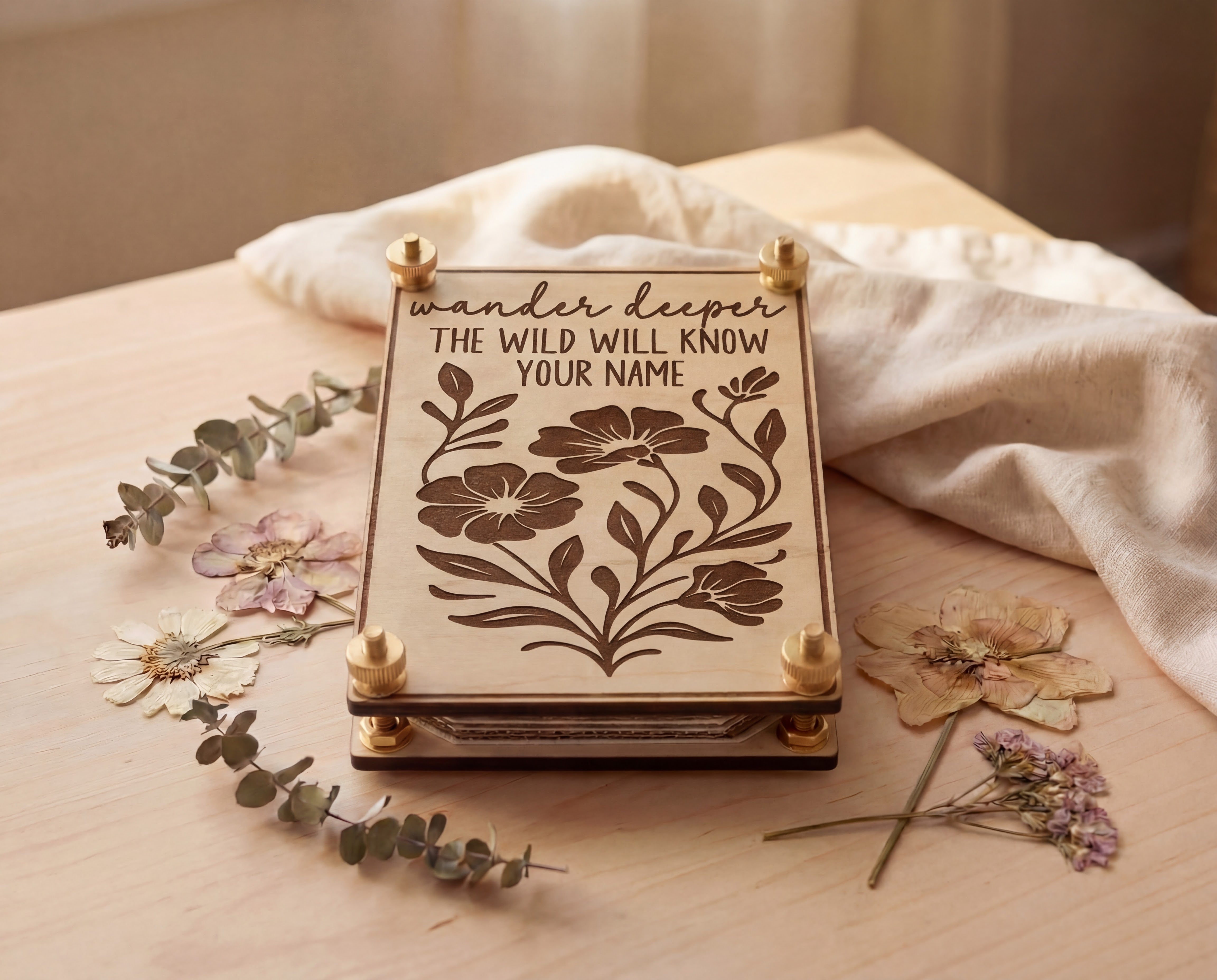 Handmade 4x6 maple flower press with brass hardware and “Wander deeper, the wild will know your name” engraved floral design, styled on a light wood table with linen cloth, eucalyptus sprigs, and pressed flowers.